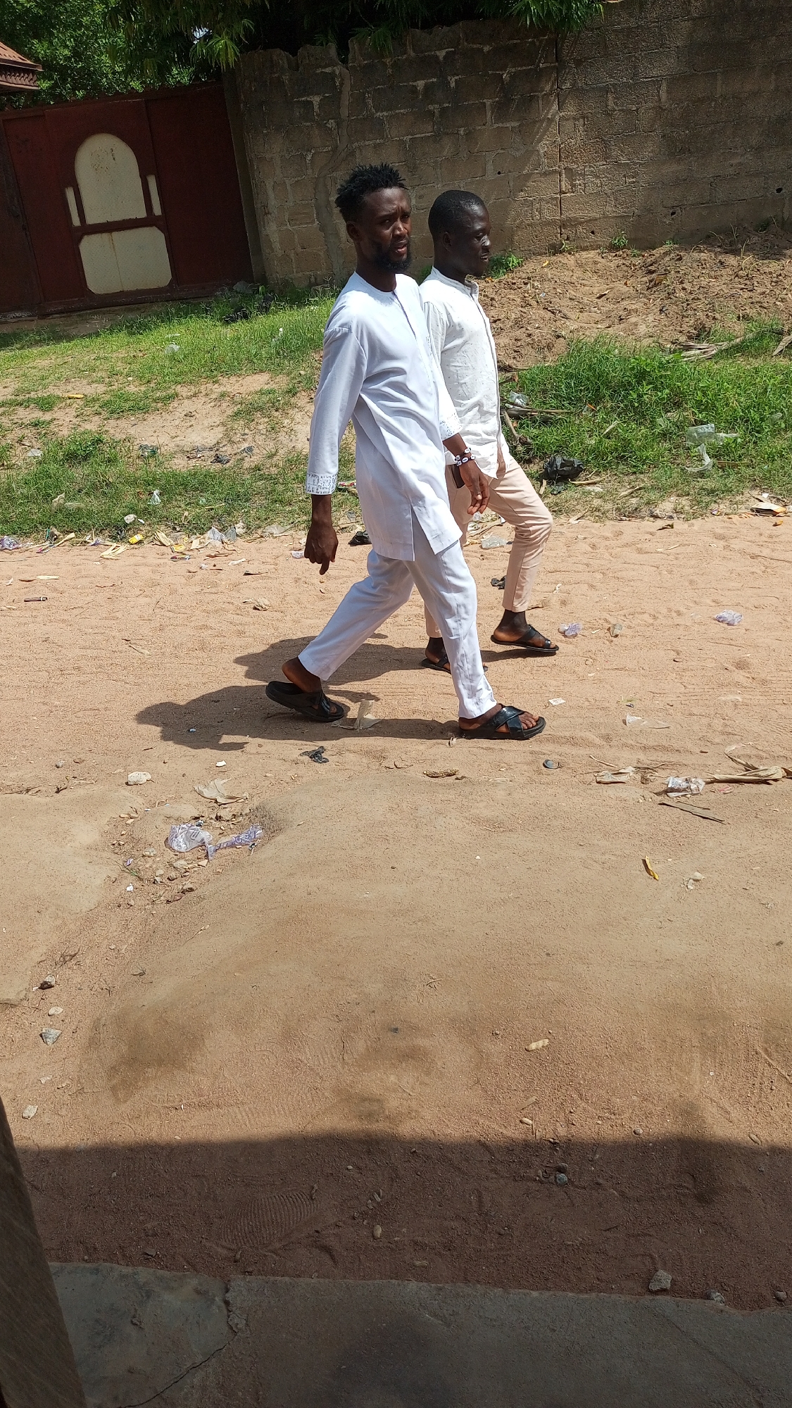 Two Black men are walking on a sandy, dirt road in Jalingo, Nigeria. The man in the foreground is wearing a white tunic and white pants with black sandals. He has short, dark hair and a beard. He is looking to his left. The man behind him is wearing a white shirt and light-colored pants with dark sandals. He has short, dark hair and is looking to his right. In the background, there is a brick wall, a large metal gate with a white inset design, and green vegetation. The ground is littered with various pieces of trash, including plastic bags and wrappers. A distinct footprint is visible in the sandy soil in the foreground, partially obscured by shadow.