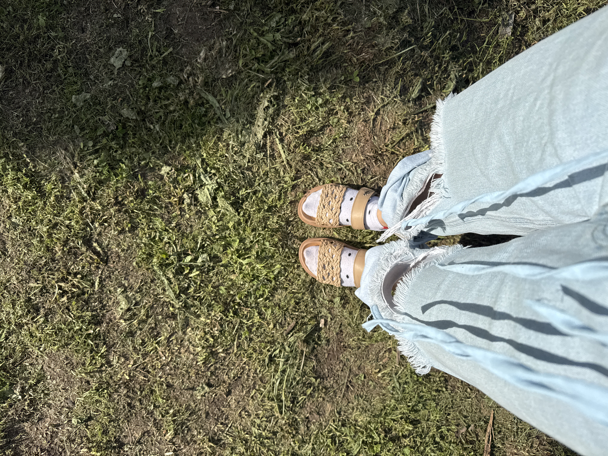 This media file captures a high-angle, first-person perspective, looking down at a person's feet and lower legs. The individual is standing on an outdoor surface composed of sparse green and dried grass mixed with visible dirt and soil, indicative of a natural, possibly unkempt, ground area.
The person is wearing light blue denim pants, which feature notably frayed and tattered hems, suggesting a distressed or casual style. Their feet are clad in sandals with light brown or tan soles and woven straps made of a natural fiber, such as rope or jute, forming an intricate pattern across the top of the foot. White socks with small black polka dots are worn with the sandals.
Strong, distinct shadows are cast across the ground by the pants and sandal straps, indicating bright sunlight and suggesting the time of day is sunny, likely mid-day or afternoon. No specific activity is depicted beyond standing or being present in this outdoor setting. There is no visible text within the image. The scene conveys a casual, outdoor ambiance, fitting for a rural or semi-rural location like Луптюг, Russia.