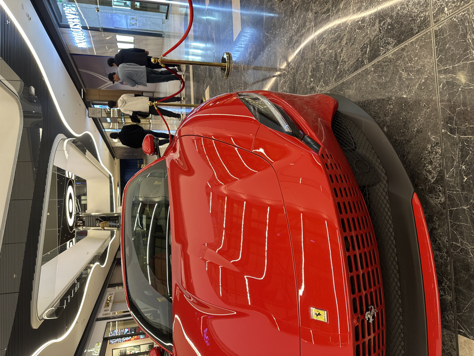 A glossy red Ferrari sportscar is prominently displayed in the foreground of a modern, multi-level indoor shopping mall in Tashkent, Uzbekistan. The vehicle, possibly a Ferrari Roma, faces slightly left of center, showcasing its front grille, headlights, and the Ferrari emblem on the hood. It is cordoned off by red velvet ropes suspended between gold-colored stanchions. The polished dark marble floor, featuring white veining, reflects the car and the mall's overhead lighting.
In the mid-ground, beyond the barrier, four adult males are observed walking or standing with their backs mostly to the camera. They are dressed in casual attire, including jackets and a hoodie. Further in the background, multiple levels of the mall are visible, characterized by curved architectural elements, linear LED strip lighting along ceilings and walkways, and several illuminated storefronts. An escalator system connects the upper levels. One storefront on the right displays the partially visible name "AVATOSLAVEE" illuminated in blue light. The overall environment is well-lit and upscale.