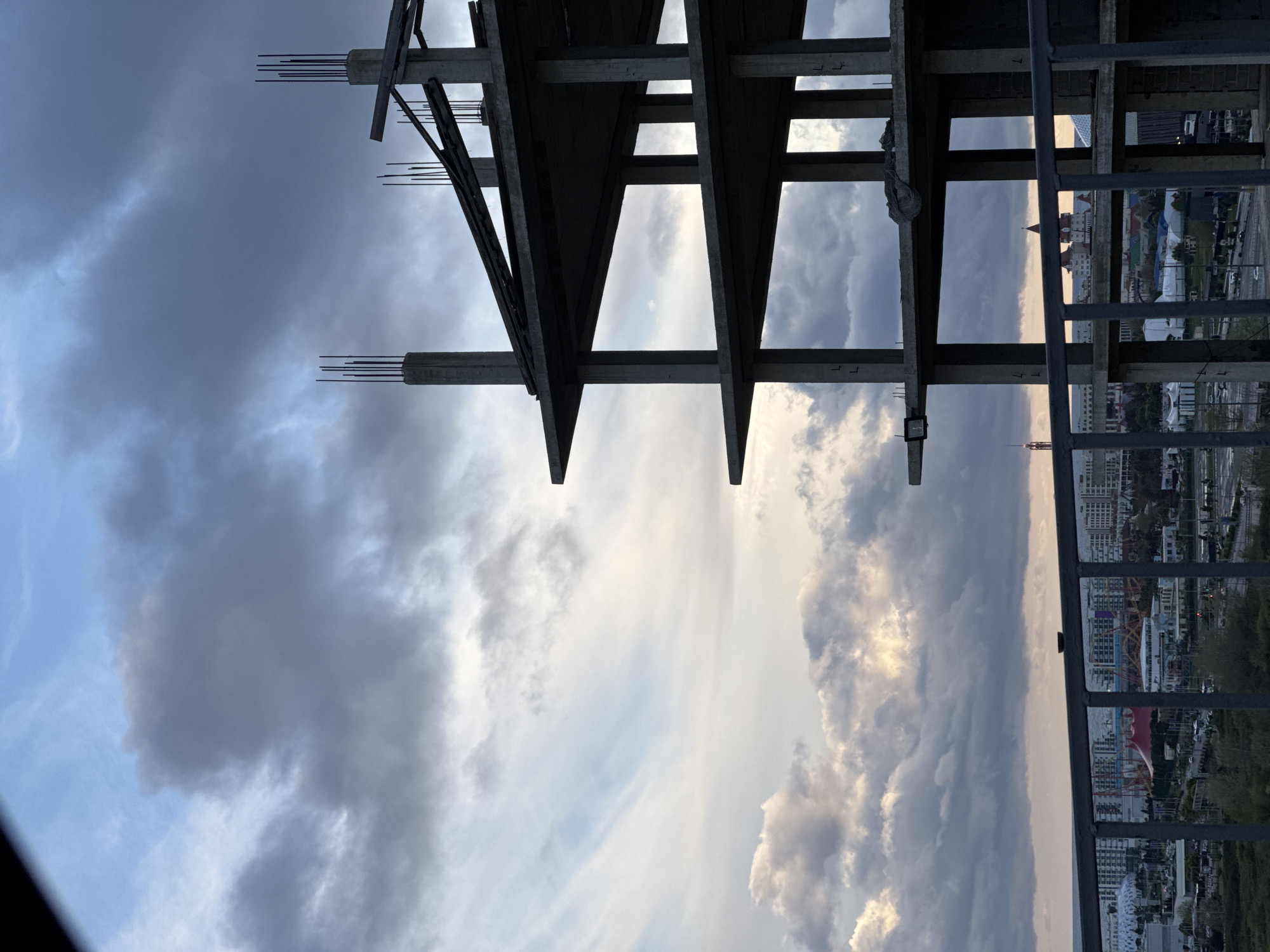 The image depicts a view from a high vantage point in Sochi, Russia, looking through the unfinished framework of a building. The foreground features the raw concrete and steel structure of the building, with protruding rebar. The background reveals a cityscape under a dramatic sky with dark, cloudy formations. A ferris wheel can be seen in the distance.