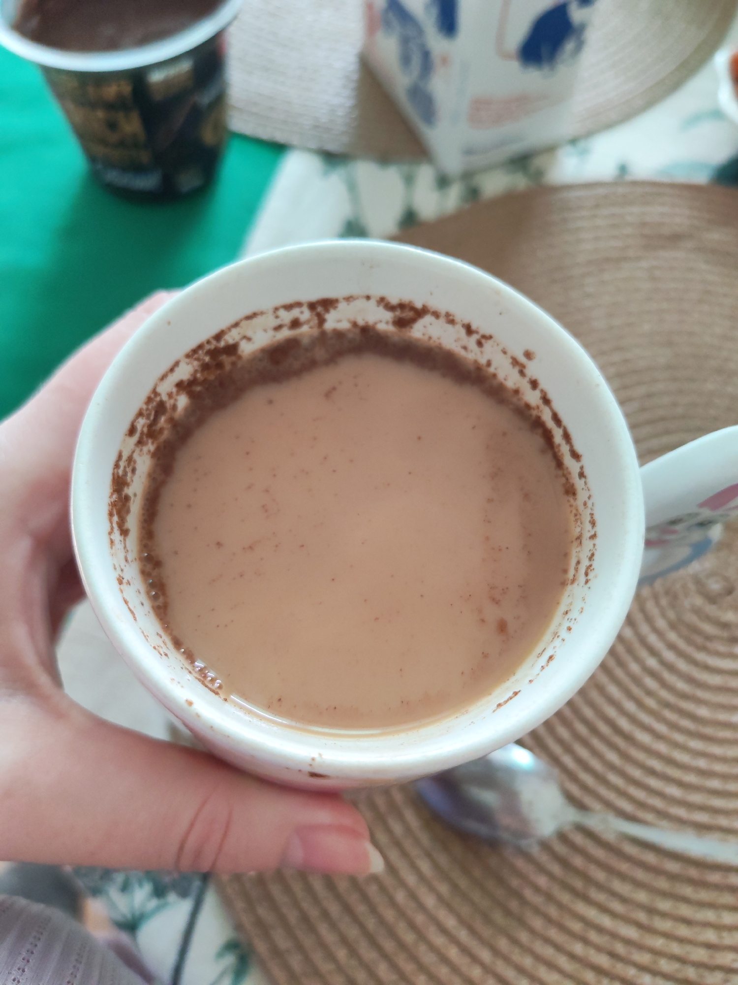 A hand is holding a white mug filled with a light brown liquid, possibly coffee with milk or tea. A brown residue is visible around the rim of the mug. A silver spoon is on a woven placemat. A container of dark-colored food, likely pudding or yogurt, is partially visible in the upper left corner. A blurry white container with blue markings appears in the background. The objects are arranged on a surface with a floral pattern in the lower left. There is no evidence to suggest this photo was taken in Izhevsk, Russia.