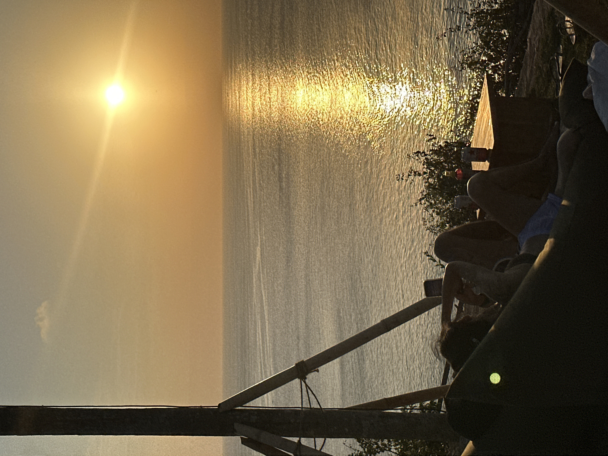 The image captures a serene sunset over the ocean, likely viewed from a clifftop or elevated vantage point in Kuta Selatan, Indonesia. The sky is painted in warm hues of orange and yellow, with the sun a bright orb just above the horizon, casting a glittering path of light across the tranquil water. In the foreground, silhouetted figures are relaxing, suggesting a moment of leisure. One person appears to be lying down, with their hands behind their head, while another is also reclined. A low wooden table is visible, holding a couple of drinks, hinting at a social gathering. Sparse vegetation dots the landscape around the seating area. To the left, a rustic wooden structure with diagonal supports is partially visible, possibly part of a viewing deck or a primitive shelter. The overall atmosphere is one of peaceful enjoyment of the natural beauty of the sunset.