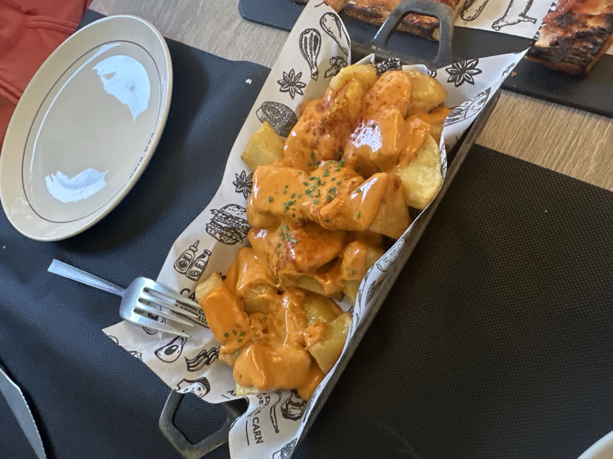 The image depicts a serving of patatas bravas, a typical Spanish dish of fried potatoes with a spicy tomato sauce. The potatoes are arranged in a metal serving dish lined with patterned paper featuring food-related graphics. They are coated in a creamy, orange-colored sauce and garnished with small green herbs. Next to the dish, on a dark gray placemat, lies a fork and a plain plate. Part of another serving dish, likely containing ribs, is visible in the upper right corner. The scene is set in what appears to be a restaurant, with a wooden table. While the city of Sant Feliu De Guíxols, Spain is mentioned in the prompt, there is no way to confirm from the picture that it was taken in this specific location.