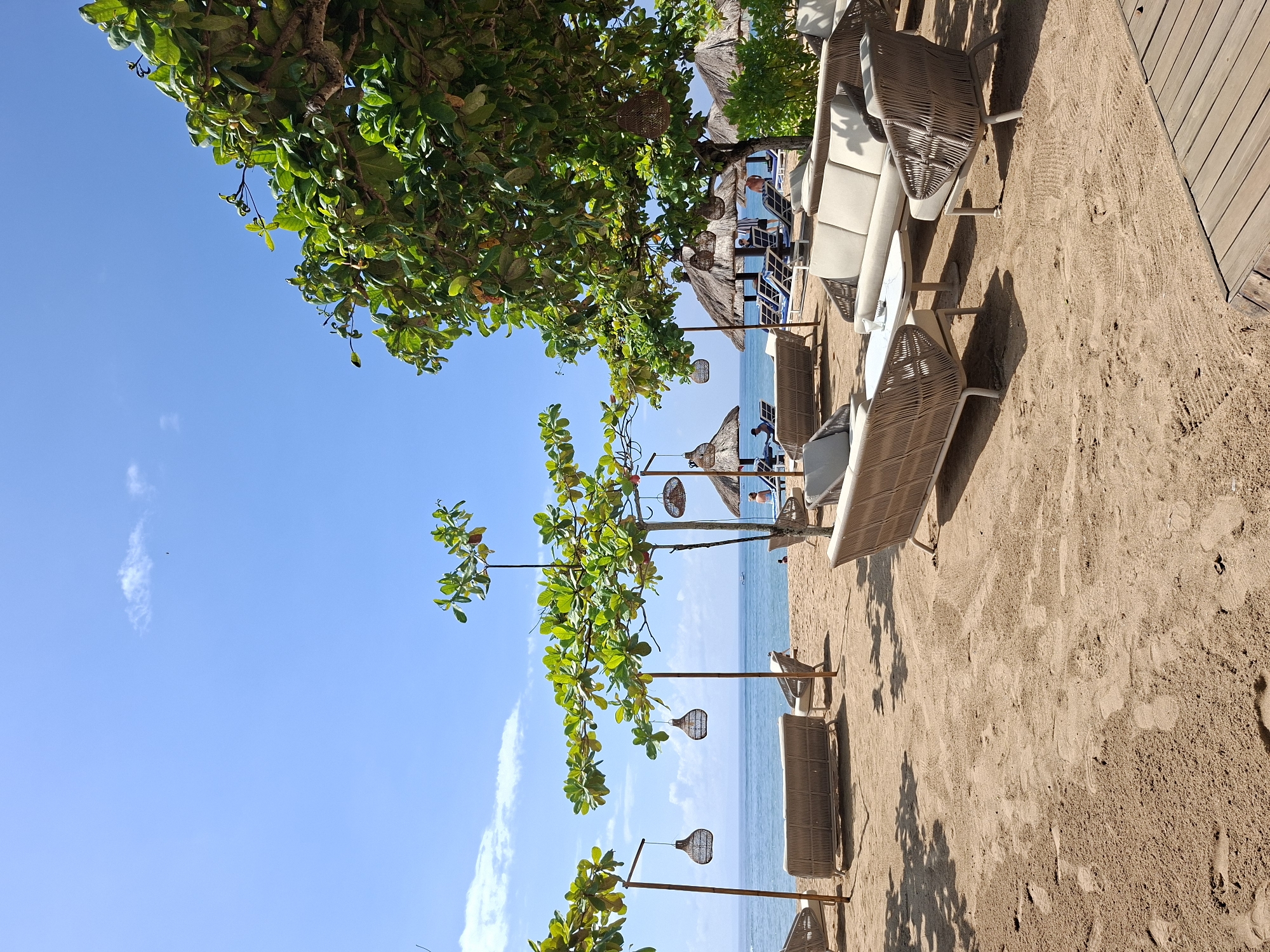 This image captures a serene beach scene at a resort in Kuta Selatan, Indonesia. The view looks towards the ocean from under the shade of lush green trees, bathed in bright daylight.
In the foreground, a sandy beach stretches across the lower portion of the frame, showing numerous footprints and textured patterns. On the sand, several modern outdoor seating arrangements are visible, including woven-style sofas and lounge chairs with light-colored cushions. Small white tables accompany some of the seating. To the far right, a section of a wooden deck or walkway is present. Strong shadows from the trees and furniture are cast on the sand, indicating a sunny day.
Further back, a calm, light blue ocean meets the horizon under a clear sky with a few wispy clouds. Closer to the water, several thatched umbrellas or hut-like structures are visible, with a few people seated beneath them, enjoying the beach. Mid-ground elements include several young trees with vibrant green leaves and multiple decorative, woven pendant lanterns hanging from poles or branches, adding to the tropical ambiance.
The overall scene conveys a sense of relaxation and leisure, typical of a tropical beach resort. There is no visible text in the image. The time of day appears to be sunny daytime, with clear weather conditions.