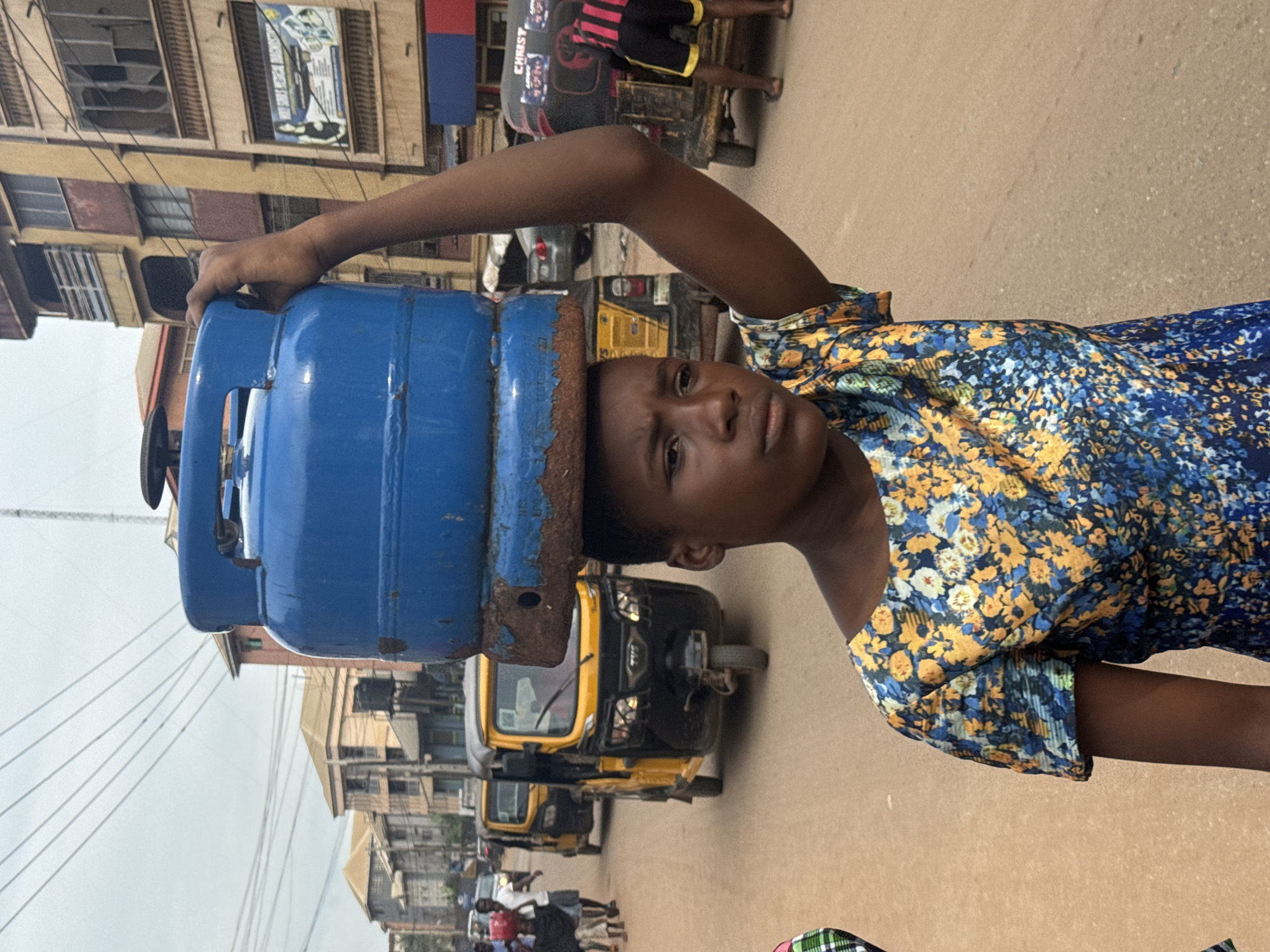 A young person carries a large blue gas cylinder on their head, balancing it with their right hand. They are wearing a short-sleeved dress with a blue, yellow, and orange floral pattern. The setting appears to be a busy street in Onitsha, Nigeria, with several yellow motorized tricycles (keke-napeps) and other vehicles visible. Buildings line the street, some appearing to be multi-story structures with balconies and windows. The sky is overcast. Other pedestrians are present in the background.
