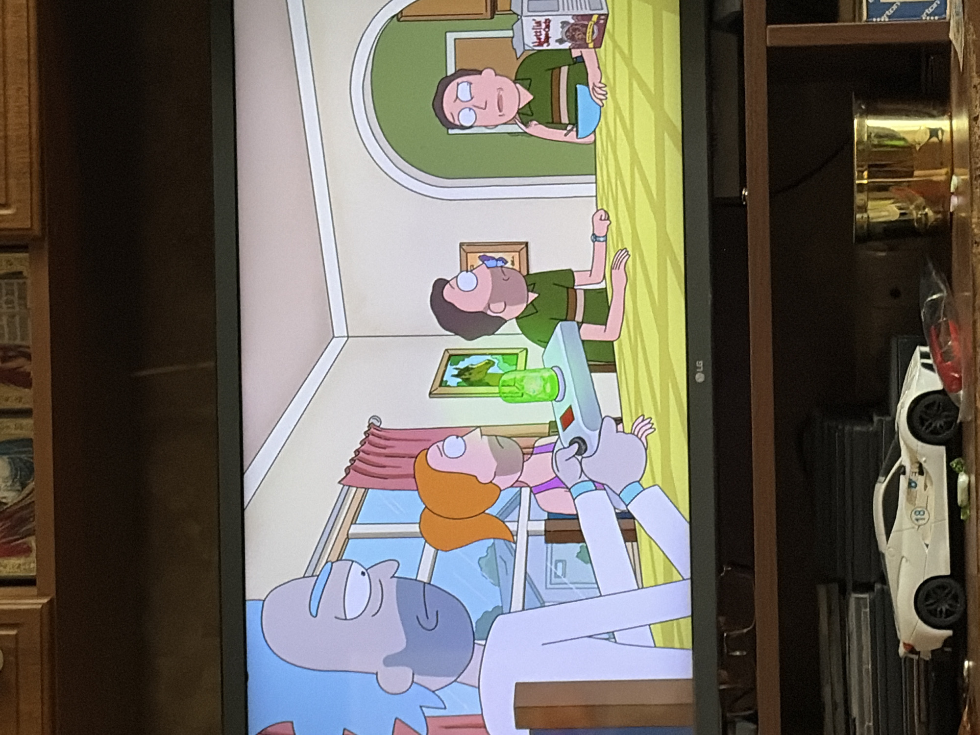 The image shows a scene from the animated television show Rick and Morty, displayed on a television screen. In the foreground, a white toy sports car is visible on a shelf below the television. The television displays an interior scene with four characters: Rick Sanchez, Morty Smith, and two other male characters. Rick is on the left, holding a device that emits a green light. Morty is to his right, wearing a purple tank top. Two men, both wearing green striped shirts, are seated at a table in the background. One of the men has a butterfly on his face. A box of cereal and a bowl are on the table. The room has yellow walls and a large window with pink curtains. The overall setting appears to be a domestic interior.