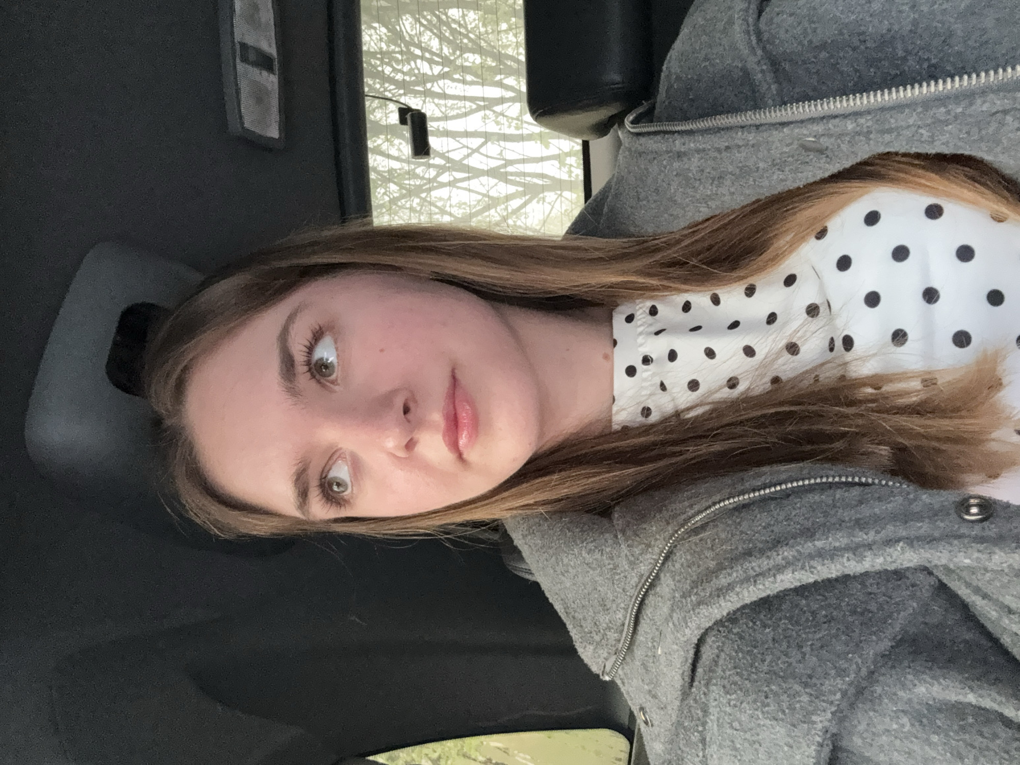 A young woman with light brown hair is seated in a car, wearing a grey hooded jacket over a white shirt with black polka dots. She is looking slightly to the left of the camera. The background includes a car window with a view of bare trees, possibly in the city of Perm, Russia, along with the car's interior, including a headrest and a ceiling light.