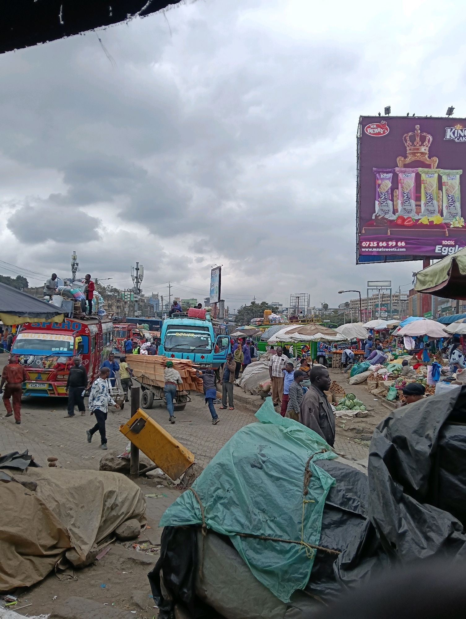 The image depicts a bustling market scene in Nairobi, Kenya, under a cloudy sky. In the foreground, several large objects are covered with tarps, one a light green and another black, with visible ropes binding them. A yellow traffic barrier lies on its side in the dirt. Beyond this, a busy street is filled with activity.
Several vehicles are present, including a red and yellow matatu with "SPERO" written on its side, a blue matatu labeled "DENGO," and another truck. People are engaged in various activities: some are standing around, others are loading and unloading goods, and a few are walking through the market.
Further back, market stalls with umbrellas and awnings are visible, with produce and goods displayed. In the distance, a large billboard advertises "MR. BERRY'S KING CAKE," featuring images of cake bars and strawberries. Communication towers and buildings are also visible in the background, indicating an urban setting. The overall impression is one of a vibrant and active marketplace.