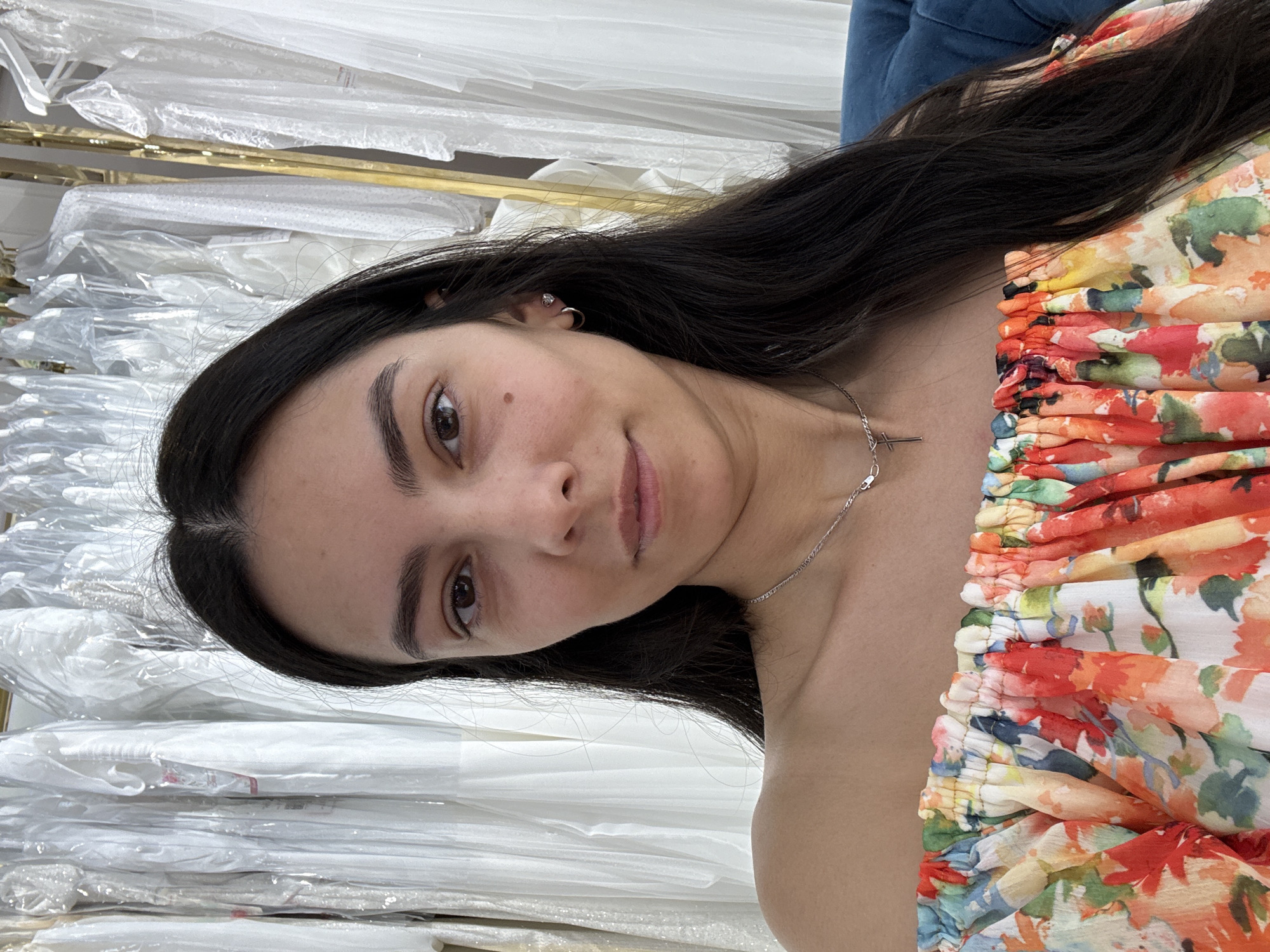 The image presents a woman in a floral off-shoulder top, with dark hair, wearing a delicate silver necklace with a cross pendant, and hoop earrings. She appears to be in a retail environment, specifically a wedding dress shop, as evidenced by the rows of white dresses, some still in protective plastic, behind her on a rack. The location is possibly in the city of Chaikovsky, Russia.