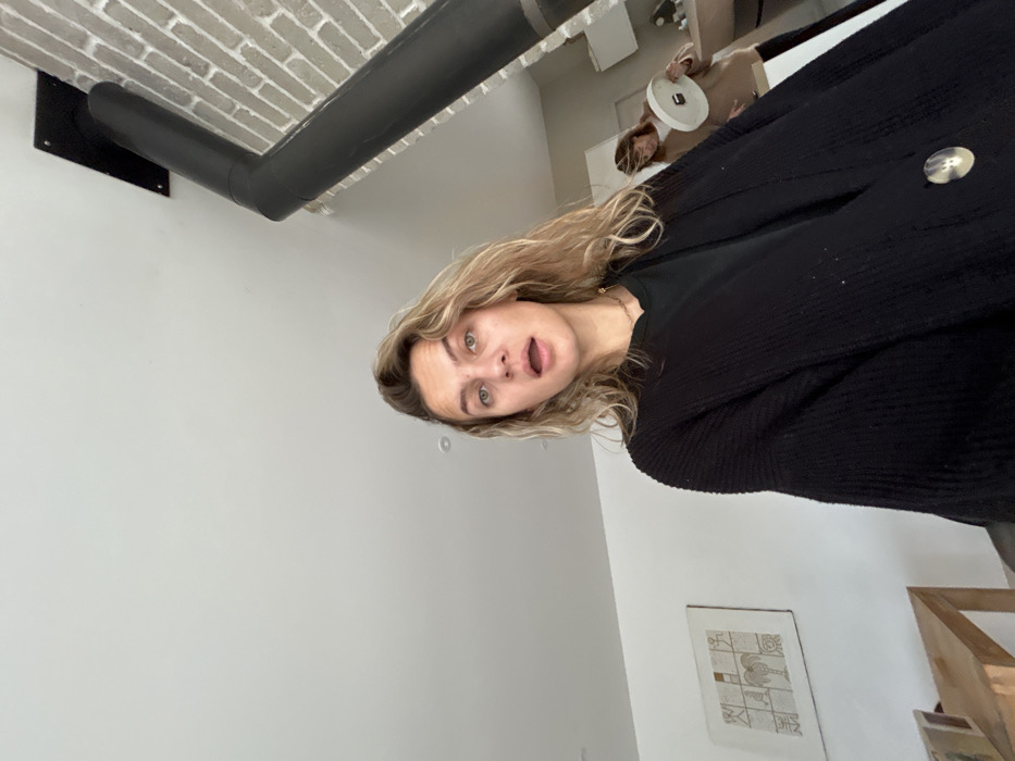 An indoor scene in Veauchette, France, features two individuals. In the immediate foreground, a light-skinned female with wavy blonde hair, green eyes, and an open mouth looks directly into the camera. She wears a black crew-neck top and a black ribbed cardigan with a single light-colored button visible on the right lapel. Her head is tilted slightly. Behind her, in the mid-ground to the right, a second individual with long dark hair, wearing a light brown long-sleeved top, is partially visible, holding a white, circular, flat object with both hands. The background consists of white walls and a white ceiling with a single recessed light fixture. A black stovepipe or chimney vent extends diagonally from a black mounting plate on a light-colored brick wall in the upper right. On the left wall, a framed artwork featuring abstract, geometric figures in earth tones is mounted. A light-colored wooden table or furniture piece is partially visible in the lower-left quadrant.