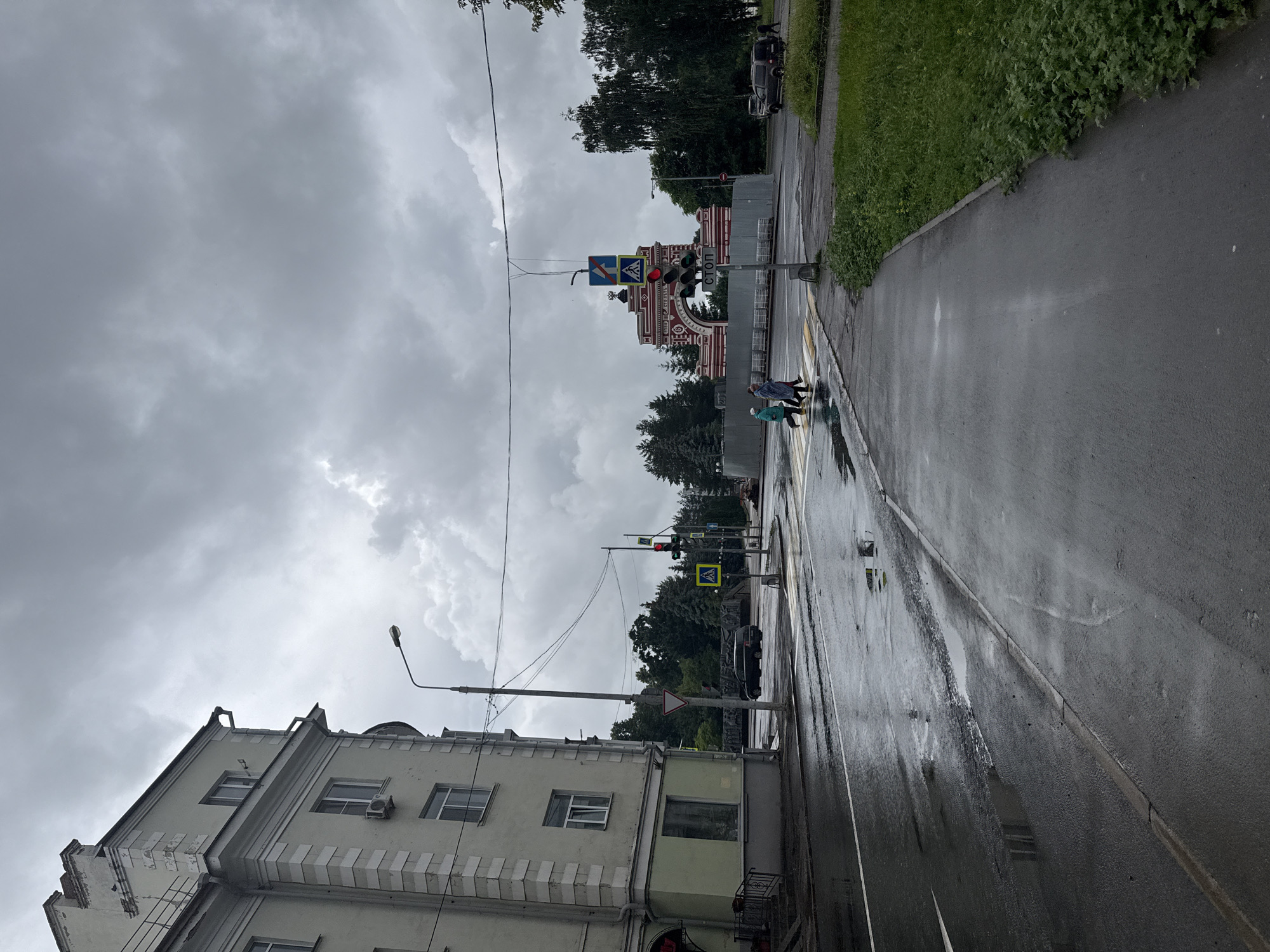 The image depicts a street scene in Kazan, Russia, under a cloudy, overcast sky, suggesting a recent or ongoing rainfall. A multi-story building with light green and white accents stands on the left side of the frame, featuring multiple windows and visible architectural details like cornices and a chimney. A street lamp is visible near the building.
The road in the foreground is wet and reflective, with puddles indicating rainfall. A pedestrian crossing with yellow and white stripes is present, and three individuals are seen crossing it. They are dressed in casual outerwear, with one person wearing a blue jacket and another in a darker coat. Two other people are walking in the distance.
Further down the street, a large, ornate archway, painted in red and white, is a prominent landmark. Traffic lights are visible near the archway, with one showing red. Various traffic signs are also present, including a blue square sign with an arrow and a white pedestrian crossing sign. A dark-colored SUV is parked on the right side of the street, and other vehicles and trees are visible in the background. Construction fencing is present around the base of the archway. A sidewalk with overgrown green grass runs along the right edge of the image. The overall impression is a city street after a rain shower.