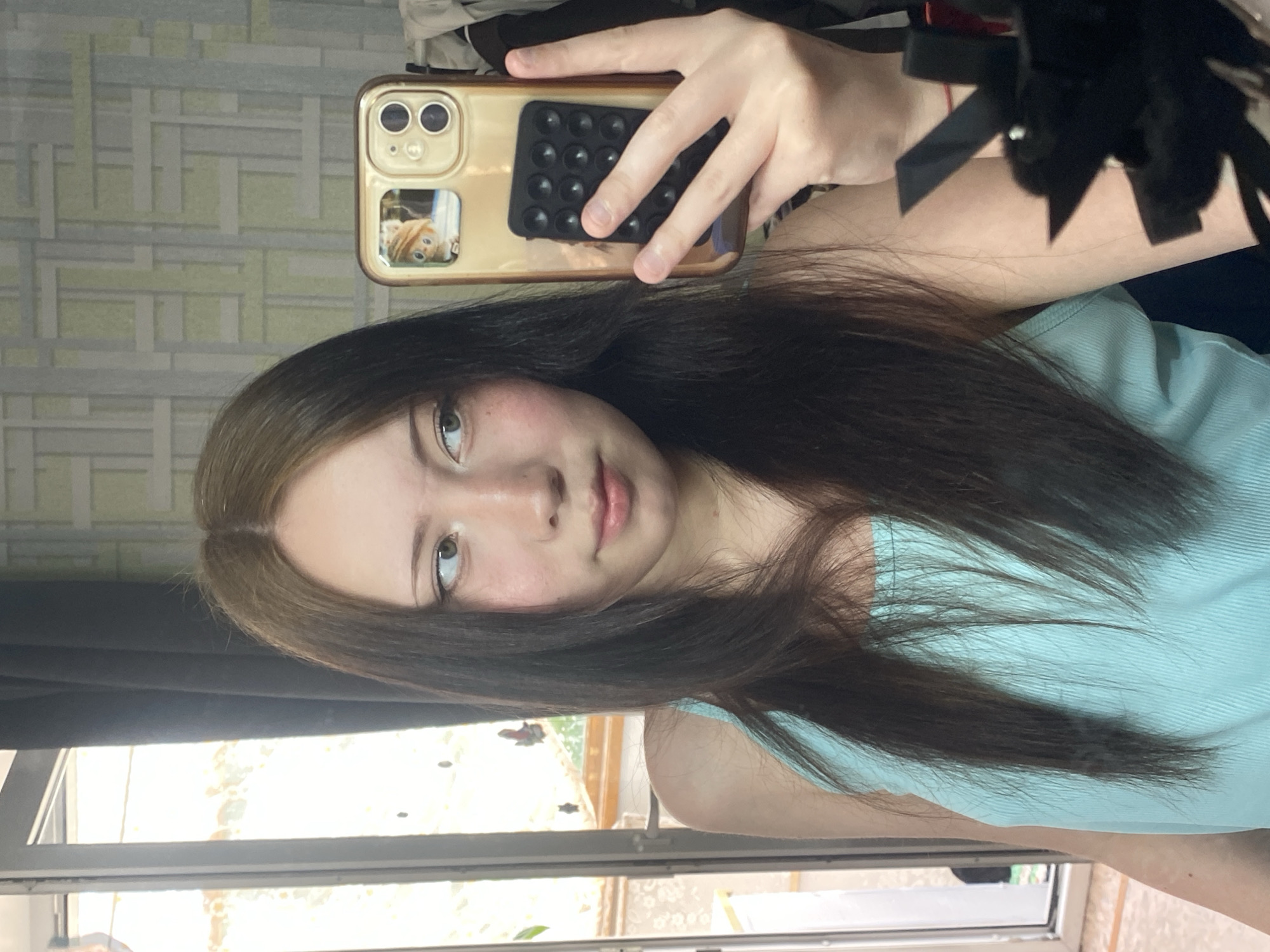 A young woman is taking a selfie indoors. She has long brown hair with lighter brown roots, light skin, and is wearing a light blue tank top. She is holding a gold-colored iPhone with a pop socket on the back, obscuring part of her face. In the background, there is a window with a view of foliage and possibly part of a building. Behind her, a wall with a geometric pattern is visible. The location is Unknown, Unknown.
