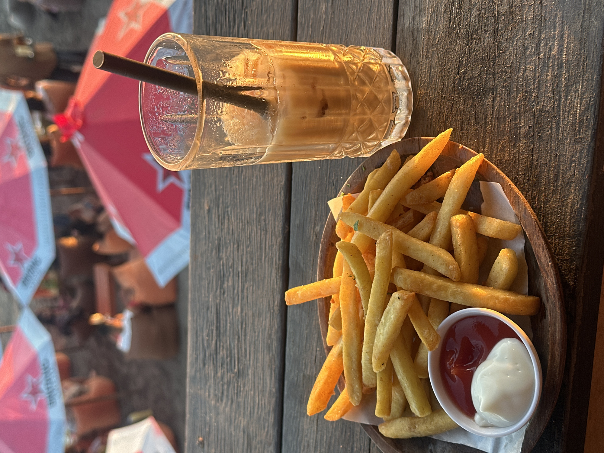 A slightly tilted medium shot shows a glass of iced coffee and a wooden bowl of french fries with a small dish of mayonnaise and ketchup on a wooden table at an outdoor location. A dark straw is in the coffee. The fries are mostly yellow. An awning with red stars and white fabric is visible in the background on the left. The location is North Kuta, Indonesia. No people are clearly visible.