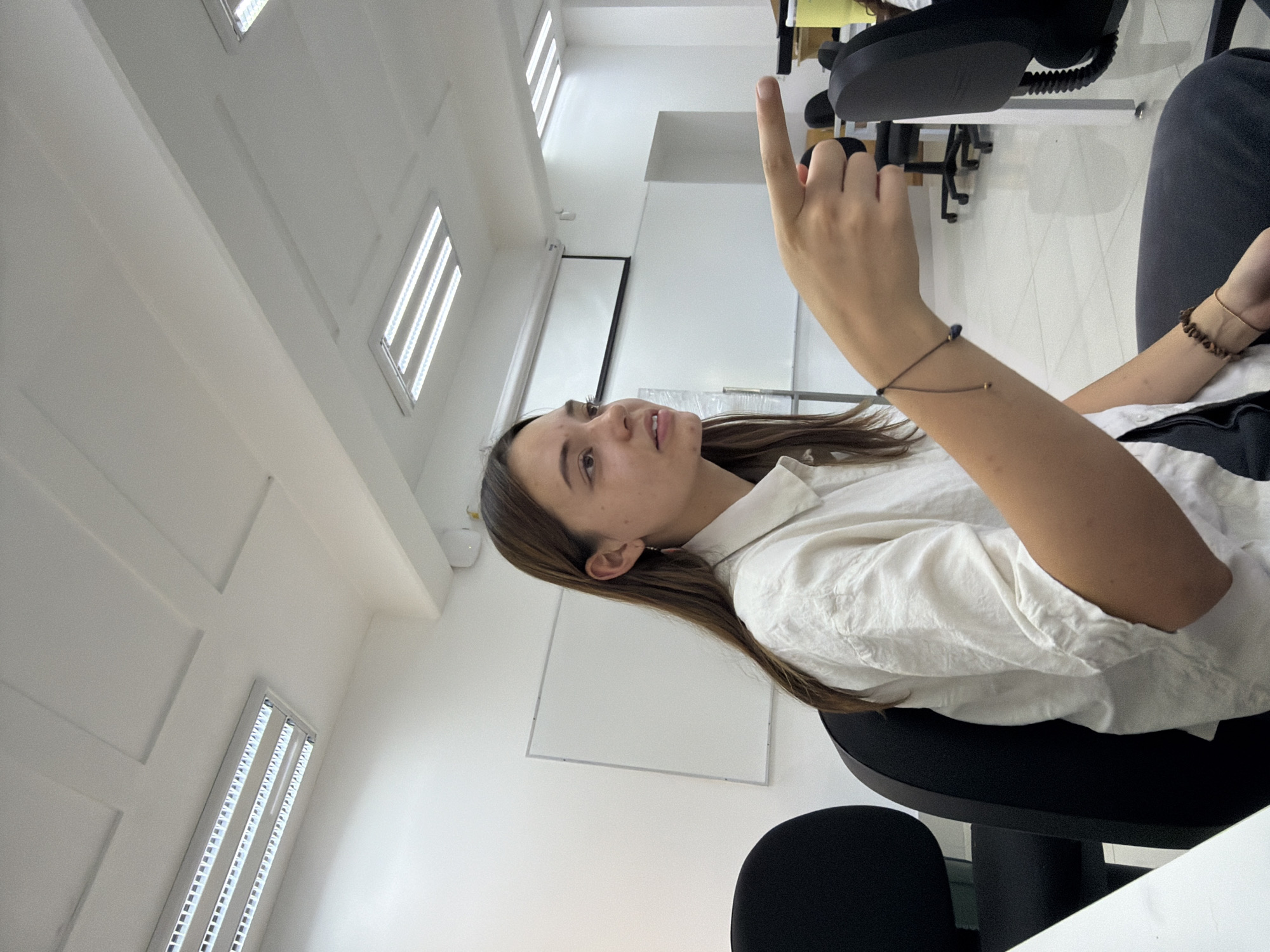 The image displays a young woman seated in a brightly lit indoor environment, likely an office, classroom, or meeting room, located in San Andrés Cholula, Mexico. The photograph is taken from a low angle, looking upwards, and is rotated, presenting the ceiling as a vertical plane on the left.
The main subject is a woman with long, straight brown hair, wearing a white long-sleeved collared shirt and dark trousers or jeans. She has delicate, simple bracelets on both wrists. Her face is in profile, looking upwards and to her left, with her mouth slightly ajar, suggesting she is attentive, engaged in thought, or about to speak. Her right arm is raised, with her index finger pointing directly upwards, as if making a point, indicating something, or asking a question.
The setting features predominantly white walls and a white ceiling, which is fitted with multiple long, rectangular fluorescent light fixtures providing bright, even illumination. These lights appear recessed within architectural panels on the ceiling. In the background, a large white projection screen or whiteboard is visible against a wall. Parts of black office chairs and a white table are also discernible, reinforcing the impression of a professional or educational space. The overall atmosphere is clean, modern, and functional. There is no visible text in the image.