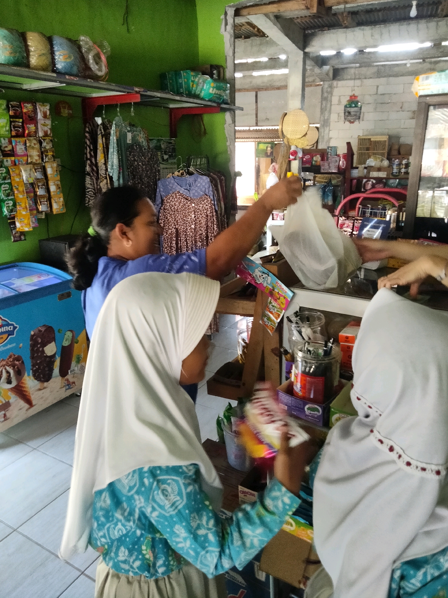 In a small shop, a woman in a blue shirt hands a plastic bag to another person wearing a white hijab and a blue and white patterned outfit. The shop appears to be a convenience store or a small grocery, with shelves stocked with various items like snacks, drinks, and clothing. An ice cream freezer is visible to the left, displaying images of ice cream cones and bars. In the background, more shelves are filled with products, and there are caged birds hanging from the ceiling. The overall environment suggests a bustling, everyday scene within a local establishment.
