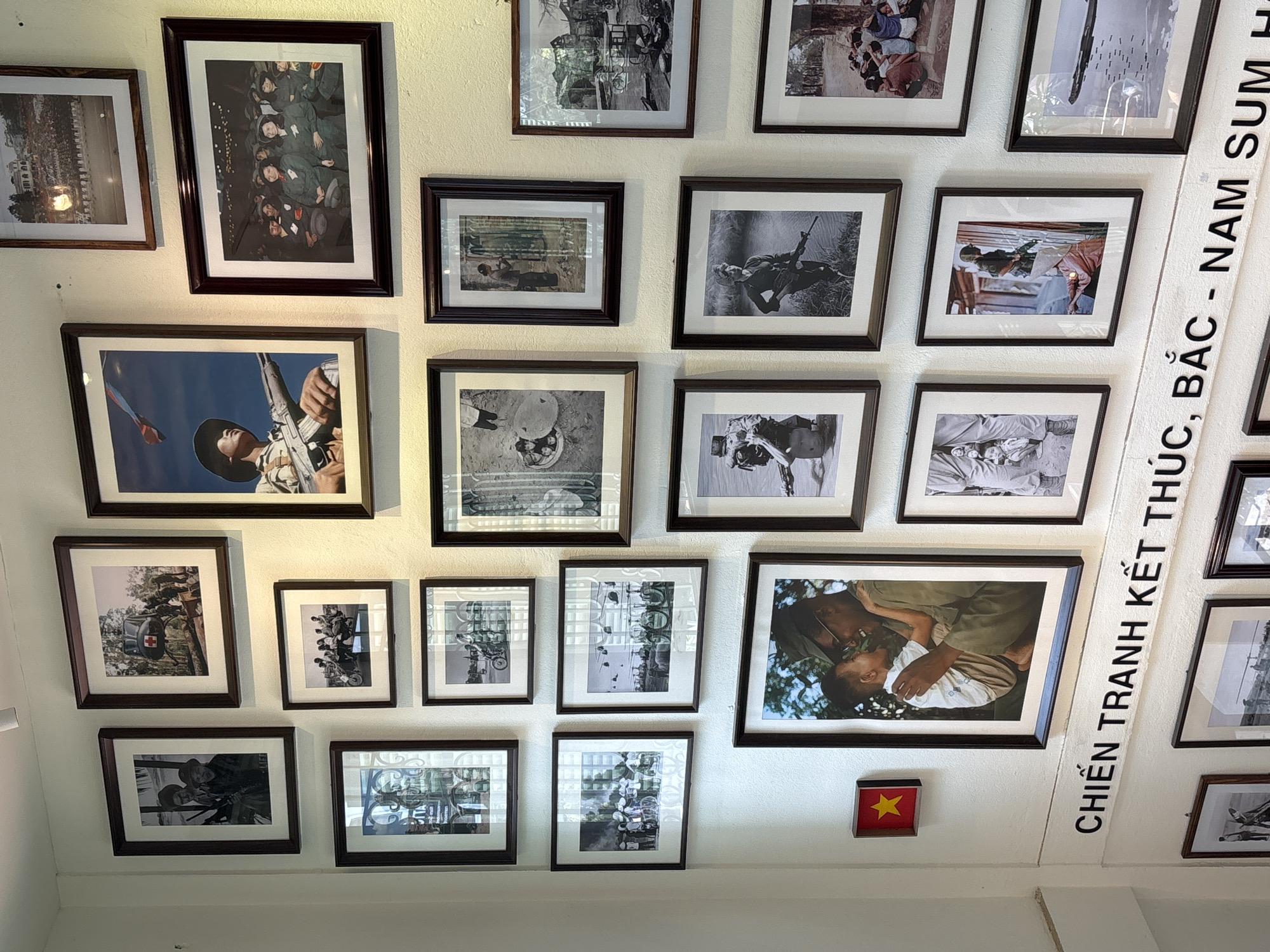 The image captures a section of a wall adorned with framed photographs, likely in a museum or exhibition space. The photographs, all framed in dark brown, depict scenes primarily related to military subjects and historical events. These images are arranged in a grid-like pattern on a white wall. In addition to the photographs, a small, square red flag with a yellow star is also mounted on the wall. A vertical inscription in Vietnamese runs along the right edge of the wall, partially obscured by the framed photos. Given the subject matter of the photographs and the location in Đà Nẵng, Vietnam, it's probable that the scene is inside a war museum or a historical exhibition related to the Vietnam War.