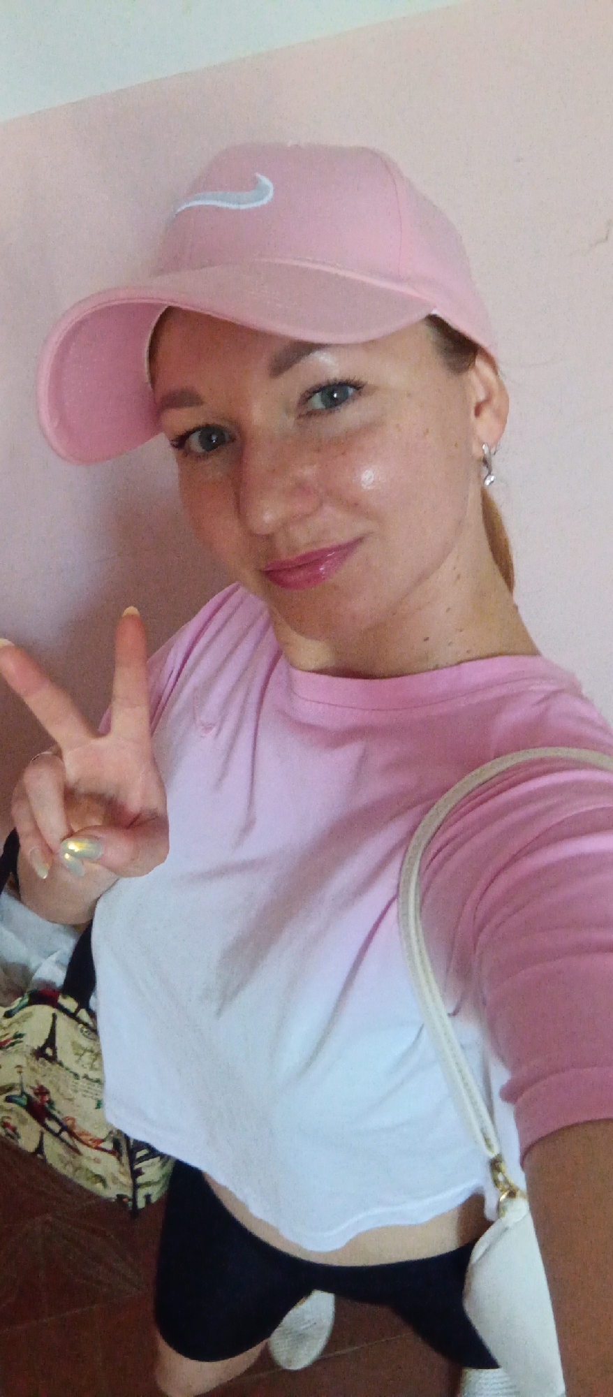 A selfie of a woman indoors. She is wearing a pink Nike baseball cap, a pink and white ombre t-shirt, and black athletic shorts. She is making a peace sign with her left hand. There is a bag with a print of Paris landmarks over her left shoulder and a small white purse hanging from her right shoulder. The background is a plain pink wall. The location in the city of Unknown, Unknown cannot be determined from the image.