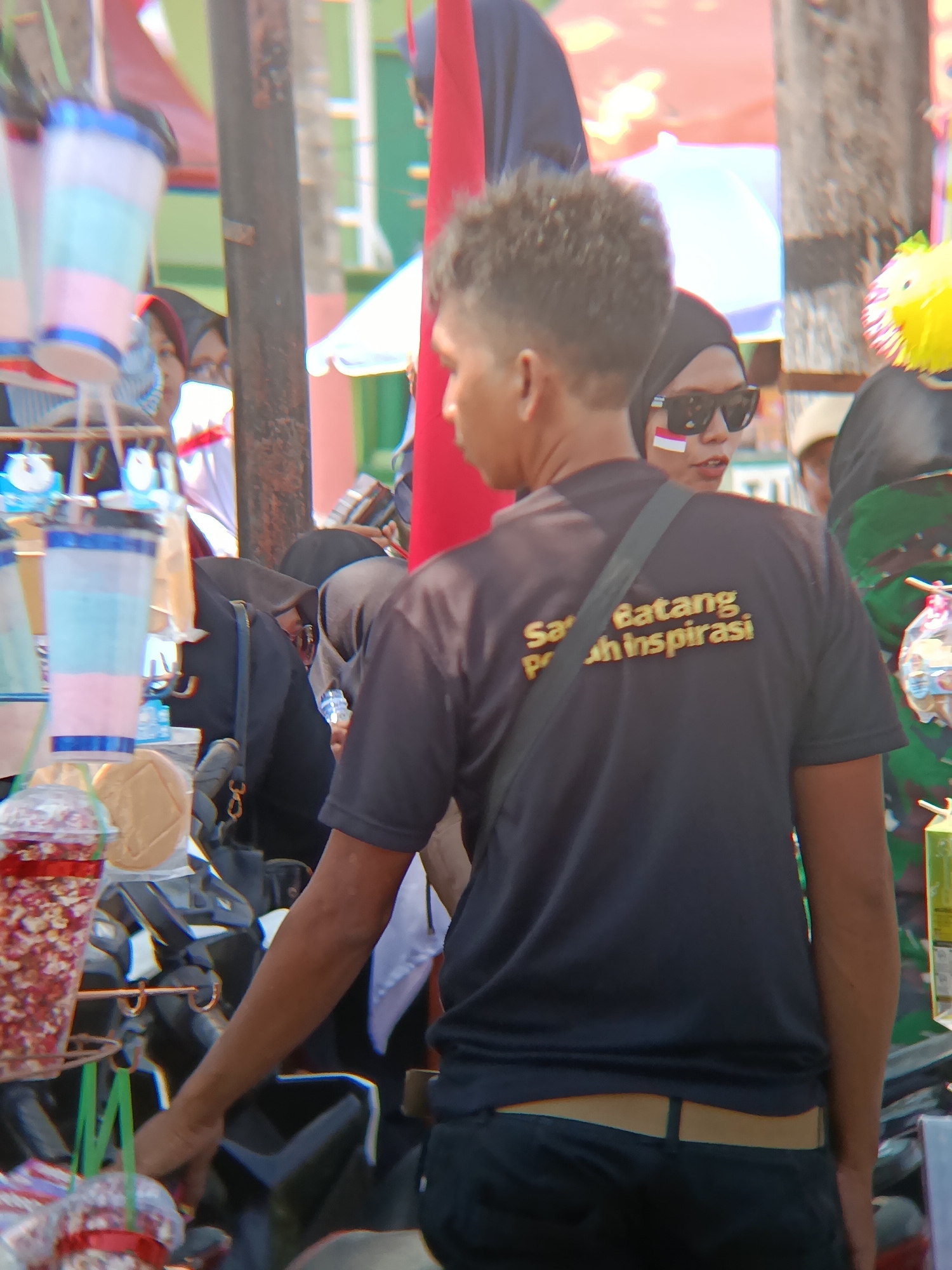 The image shows a crowded outdoor scene, possibly a street market or festival, in Desa Kebon Manis, Kecamatan Cilacap Utara, Indonesia. In the foreground, a person wearing a black t-shirt with gold lettering that reads "Sarat Batang Pohon Inspirasi" is visible from the back, with a strap from a bag across their shoulder. The person is wearing black pants with a beige belt. To the left, there are colorful plastic cups hanging, and to the right, a person wearing a black headscarf and sunglasses with a small Indonesian flag sticker on their cheek is visible. Other people and various stalls or displays are present in the background, suggesting a bustling environment. The lighting suggests it is daytime, with a bright, possibly sunny, atmosphere.