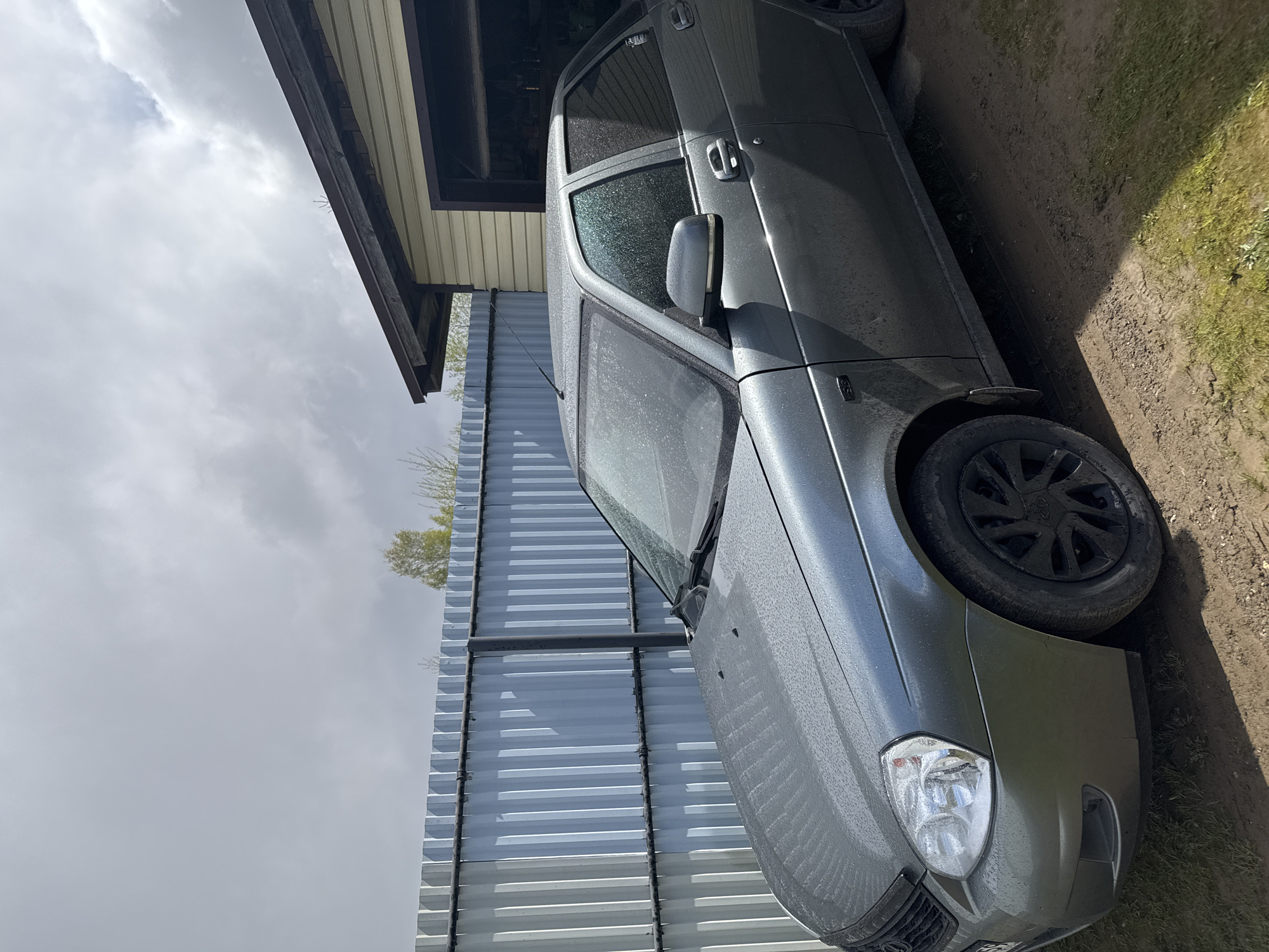 A gray Lada Priora is parked next to a galvanized steel fence and a building with siding. The ground is dirt and grass. The sky is cloudy. The location is likely in Бородулино, Russia.
