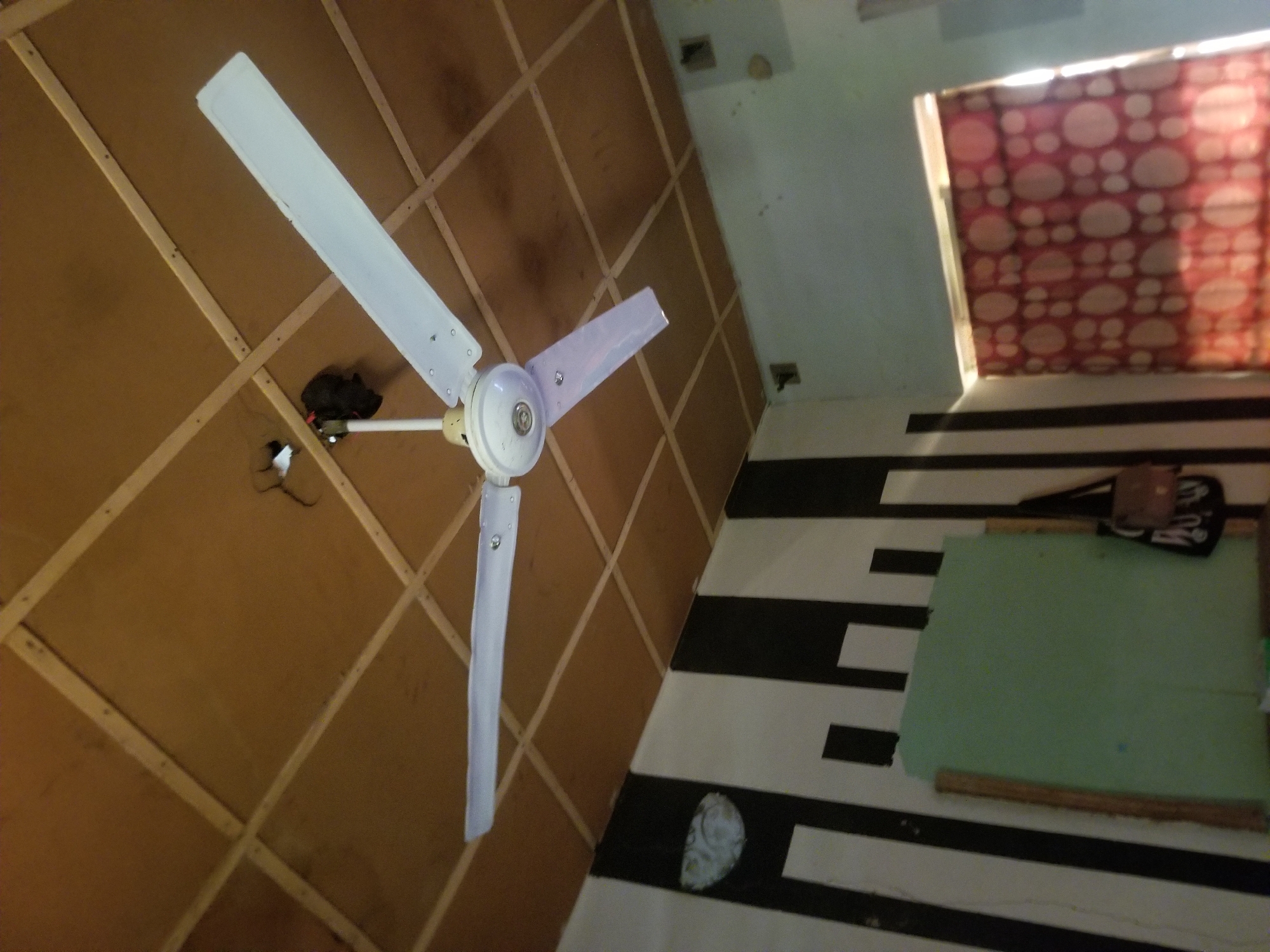 A white ceiling fan with three blades is mounted on a wooden panel ceiling. The fan's central housing is white, and the blades are white with silver screws. Above the fan, on the ceiling, a dark, possibly fabric item is attached. The walls feature a pattern of alternating black and white vertical stripes. To the left, a light green board is propped against the striped wall, and a brown purse hangs next to it. To the right, a window is covered by a red curtain with a white circular pattern. The room appears to be dimly lit, with light entering from the window. The scene is a room in Jauro Yinu, Nigeria.