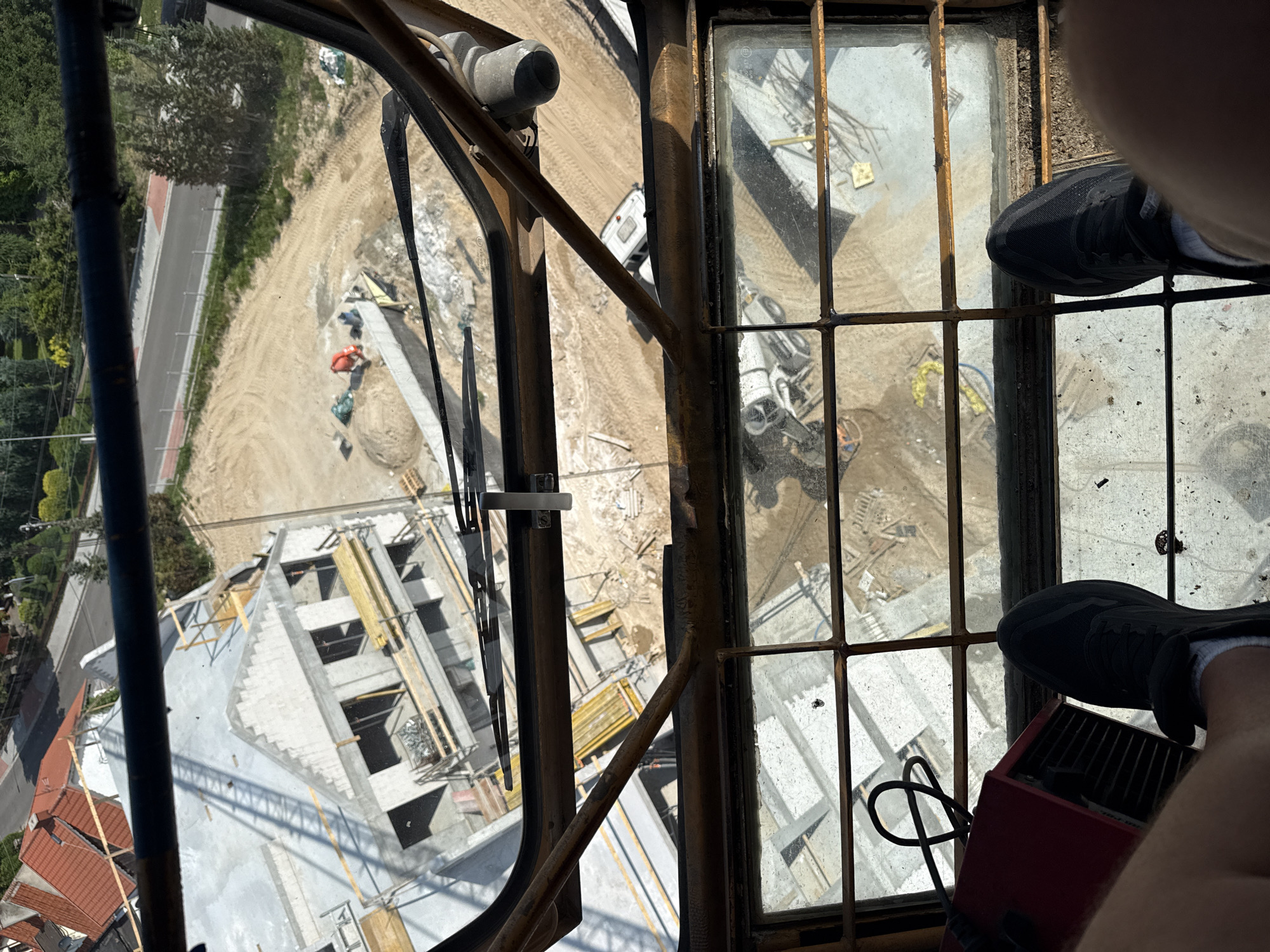 The image captures a view from a construction crane cabin overlooking a residential construction site in Mikołajki, Poland. In the foreground, a person's feet wearing black athletic shoes are visible, resting on the cabin floor. To the left, a red electrical device with a black cord is situated. The cabin features a window with a metal grid, through which the construction site and surrounding environment are viewed.
The construction site below shows a partially built house with visible concrete elements, scaffolding, and construction materials. Further back, a dirt area is present, with mounds of sand and construction equipment, including what appears to be a concrete mixer. A white vehicle, possibly a camper or small truck, is parked on the site. Beyond the construction area, a road is visible, lined with trees and residential buildings with red-tiled roofs, consistent with the architectural style of Mikołajki. The overall scene depicts an active construction project in a suburban or semi-rural setting.