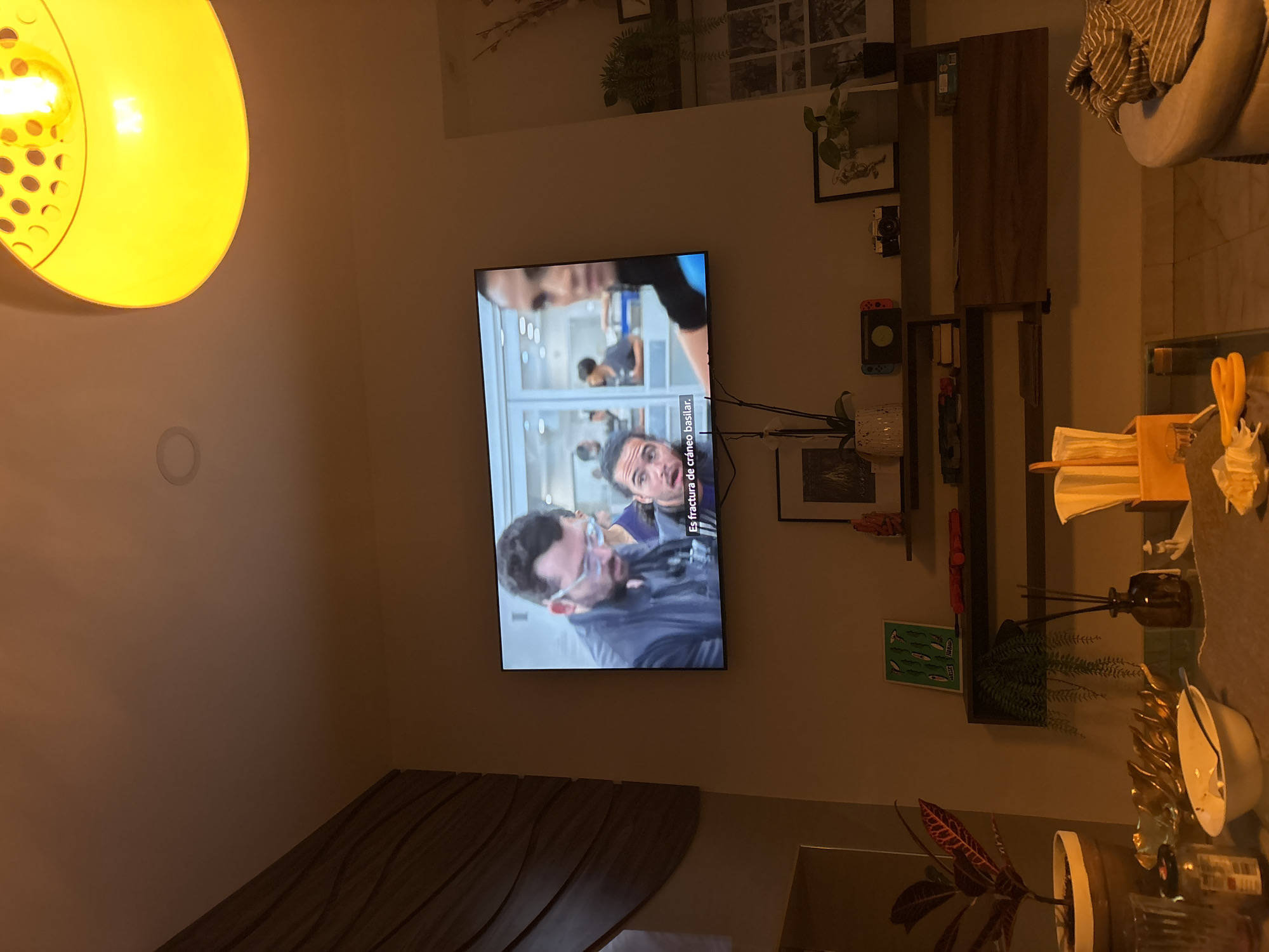 A low-angle, dimly lit interior shot shows a wall with a television screen displaying a scene with two men, one wearing glasses and a blue shirt. Text in Spanish is visible on the screen. Above the TV are decorative items on a shelf, including plants and framed pictures. To the side of the TV is a dark wooden shelving unit with various objects, including a Nintendo Switch, plants, and a small wooden box with papers and utensils. In the foreground, there is a table with a white bowl, decorative golden items, and other objects, including a glass bottle, arranged on top. The lighting in the room is provided by a large, circular yellow lamp hanging in the upper left corner. The photograph could be in a residential area, perhaps near a residential area of Puebla, Mexico.