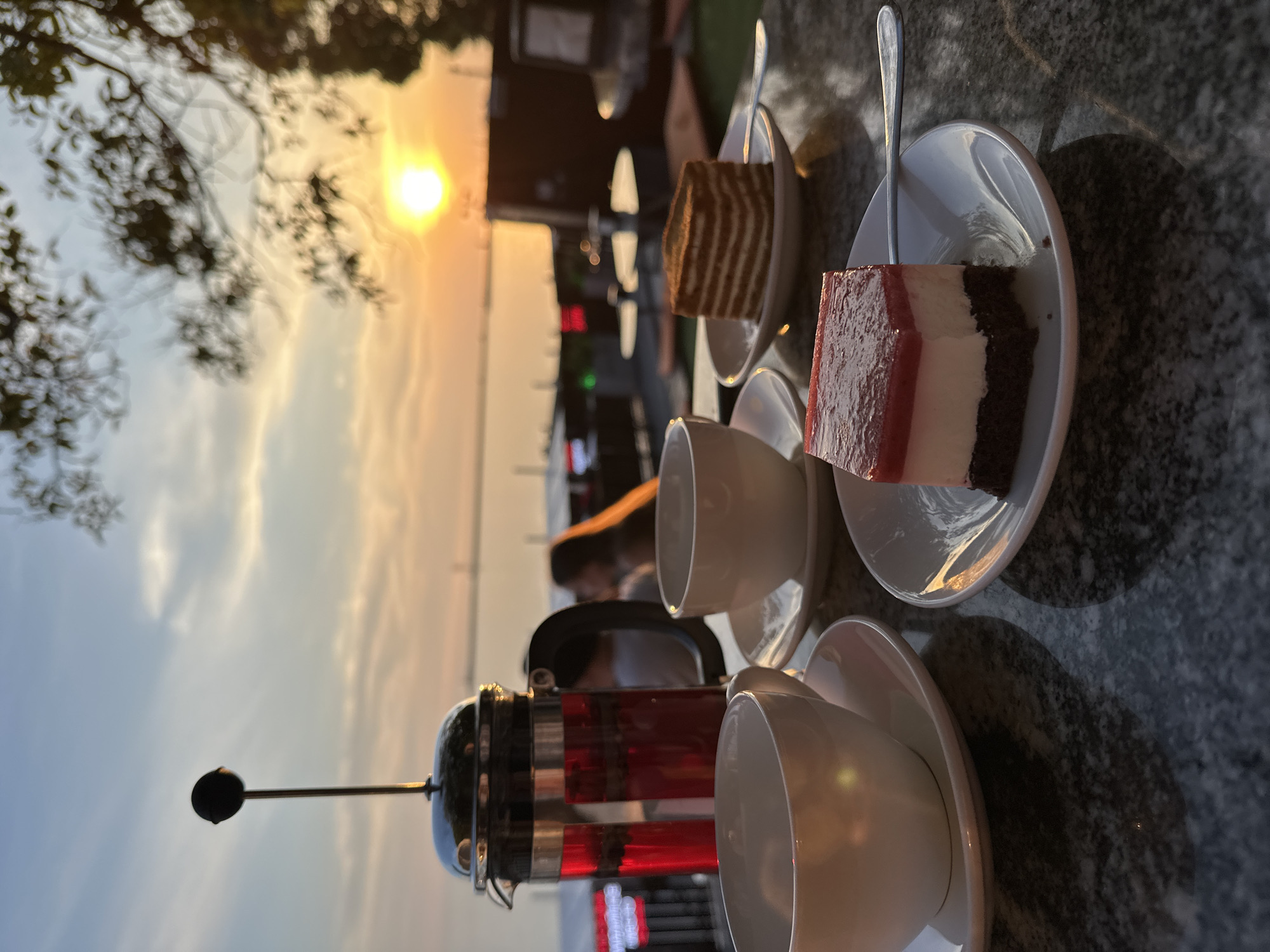 The image displays an outdoor cafe setting in Sochi, Russia, at sunset. In the foreground, a dark, mottled granite table features two white ceramic teacups with saucers. A glass French press, filled with a vibrant red liquid suggesting herbal tea, stands behind the leftmost cup. Two dessert plates are presented: the one on the left holds a rectangular multi-layered cake with a dark brown base, a white cream layer, and a red gelatinous top, accompanied by a silver spoon. The plate on the right presents a round, horizontally layered cake, also with a silver spoon. Reflections of the setting sun are visible on the polished surfaces of the cups, saucers, and spoons. The midground is out of focus, showing blurred figures of patrons, including a person with long, light-colored hair illuminated by the sunlight, and distant red illuminated signs. The background is dominated by a clear sunset over a large body of water, identifiable as the Black Sea. The sky transitions from light blue at the top to bright orange and yellow near the horizon, with the sun appearing as an intense yellow disc. Silhouetted tree branches and leaves partially frame the upper right corner of the scene, and a faint structure, possibly a bridge, is discernible in the distant horizon.