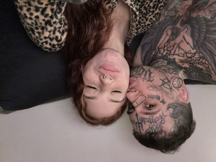 The image displays two individuals, a male and a female, in an indoor setting. The male, on the left, has short, dark hair and extensive black tattoos covering his face, neck, and upper torso. Facial tattoos include stylized script and a trident symbol on his forehead, Sanskrit/Devanagari script below his right eye, an anarchy symbol on his left cheek, and a band-aid sticker on his right cheek. He wears a large ear gauge in his left earlobe. His chest and neck exhibit intricate patterns and a large bird tattoo. He is wearing a dark, plain shirt. The male is shown puckering his lips and kissing the female's right cheek.
The female, on the right, has long, wavy, reddish-brown hair. Her eyes are closed, and her lips are puckered in a "duck face" expression. She has a silver hoop piercing in her septum. She is wearing a leopard-print garment. Both individuals appear to be seated or reclining against a dark, soft surface, which could be a couch or bed. A plain, light-colored wall is visible in the upper background. The lighting is soft and ambient. There are no visual cues within the image to determine its specific location in Buenos Aires, Argentina.