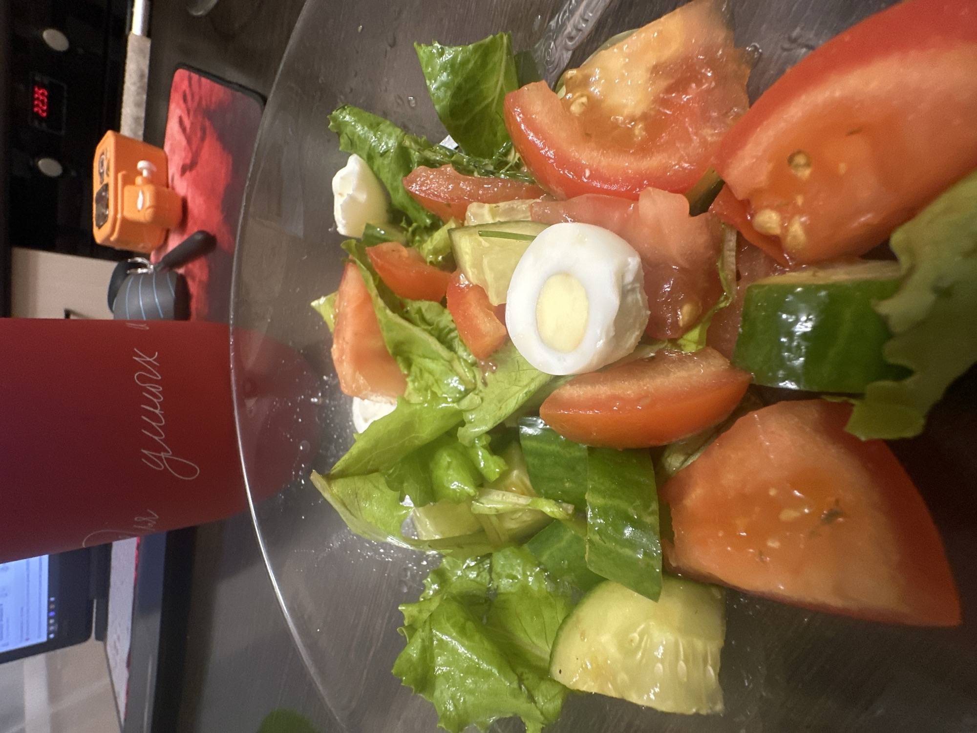 A close-up, overhead shot shows a fresh salad in a clear bowl. The salad contains chopped tomatoes, lettuce, cucumber slices, and pieces of white cheese. A halved hard-boiled quail egg sits prominently in the center of the salad. The ingredients appear moist and recently prepared.
In the background, a red cylindrical object with white cursive writing is visible on the left side of the frame. Next to it, an orange device with buttons and a small screen is placed on a red surface. A digital display showing "2235" is also visible. The overall setting suggests an indoor environment, possibly a kitchen or dining area. The lighting is dim, indicating it could be evening or a poorly lit room. The image's orientation is slightly tilted. There are no people visible in the frame.