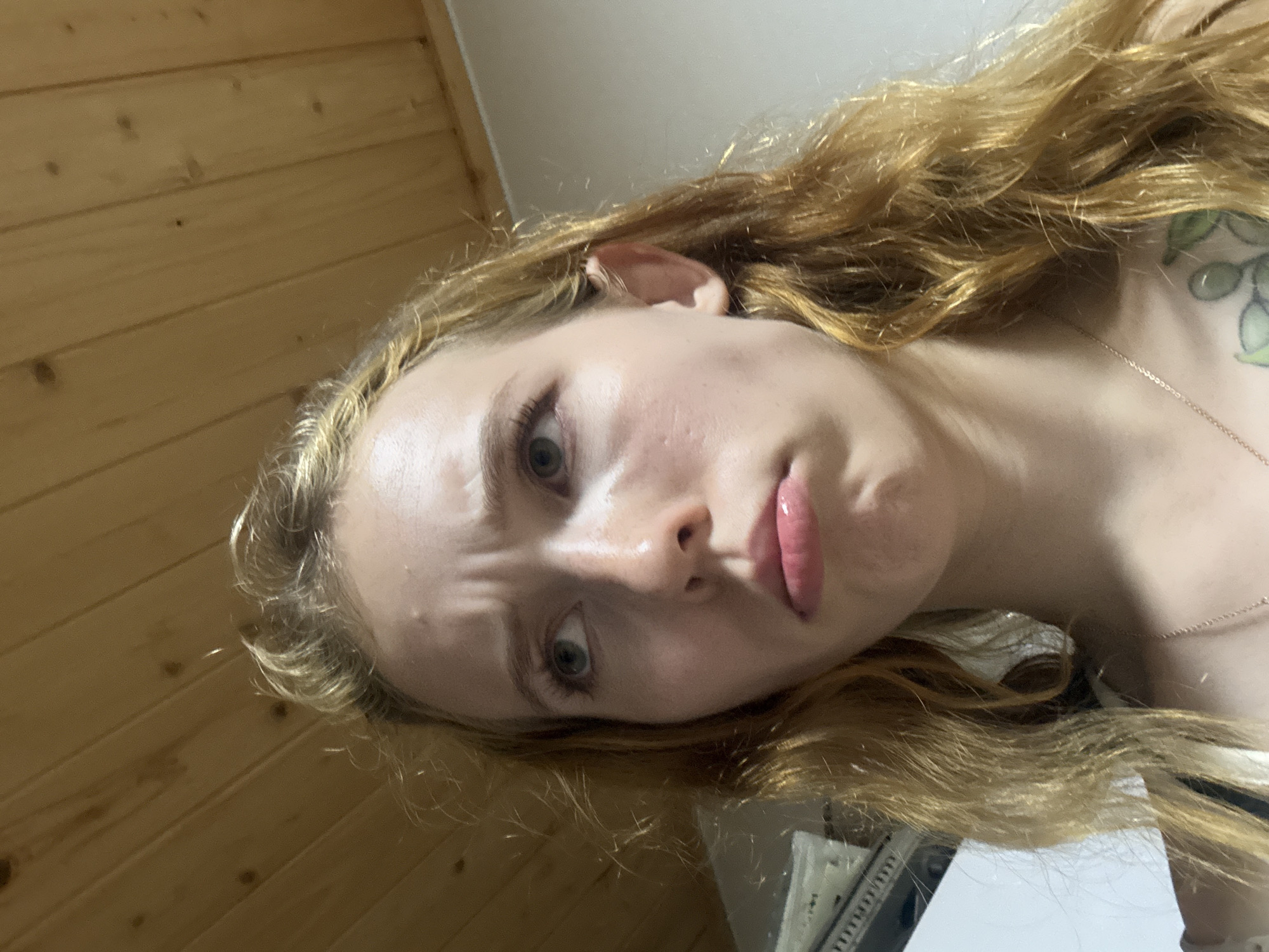 A young woman with long, wavy blonde hair is looking upwards and to her left with a pouty expression. She has light skin, green eyes, and is wearing a necklace. The background consists of a light-colored wall and a wooden paneled ceiling. The lighting suggests an indoor setting, potentially a room. There is no information provided about the location in Gwangju, South Korea.