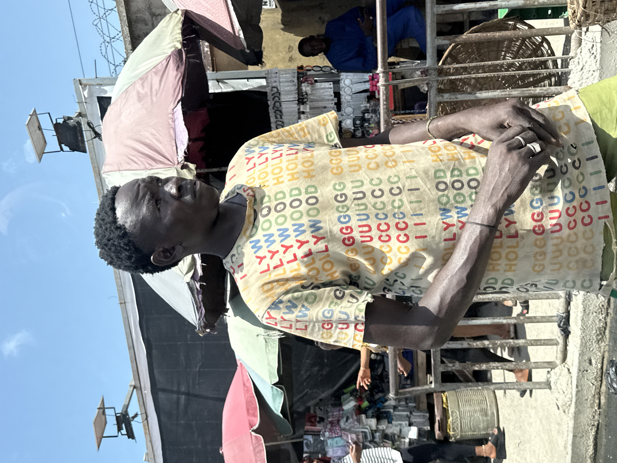 A man with dark skin and short, dark hair is centered in the frame, facing left. He wears a tan shirt patterned with colorful "HOLLYWOOD" lettering. His hands are clasped together, adorned with rings and a bracelet. In the background, a market scene unfolds under makeshift tents with colorful canopies, suggesting an outdoor vendor area. Merchandise like metal trinkets are visible on tables. Other people are partially visible in the periphery, including a person in a blue shirt seated in the background. The sky is blue in the upper left corner. The location is likely Lagos, Nigeria, evidenced by the urban market setting and local attire.
