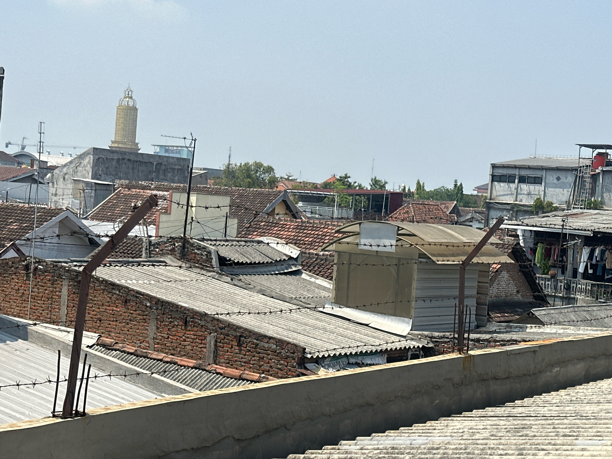 The image depicts a cityscape viewed from a rooftop, likely in Purwotengah, Indonesia. The foreground features a concrete barrier with corrugated metal roofing beyond it. Several layers of rooftops, constructed from corrugated metal and red tiles, are visible. Barbed wire fences crisscross the scene, suggesting a secure or restricted area. Buildings vary in style and condition, with some appearing to be houses and others more industrial or commercial. Among the structures, a prominent golden tower with a crown-like top stands in the distance, suggesting it might be a religious or historical landmark. The sky is clear and bright, indicating daytime with good weather. The overall impression is one of dense urban development with a mix of older and newer construction.