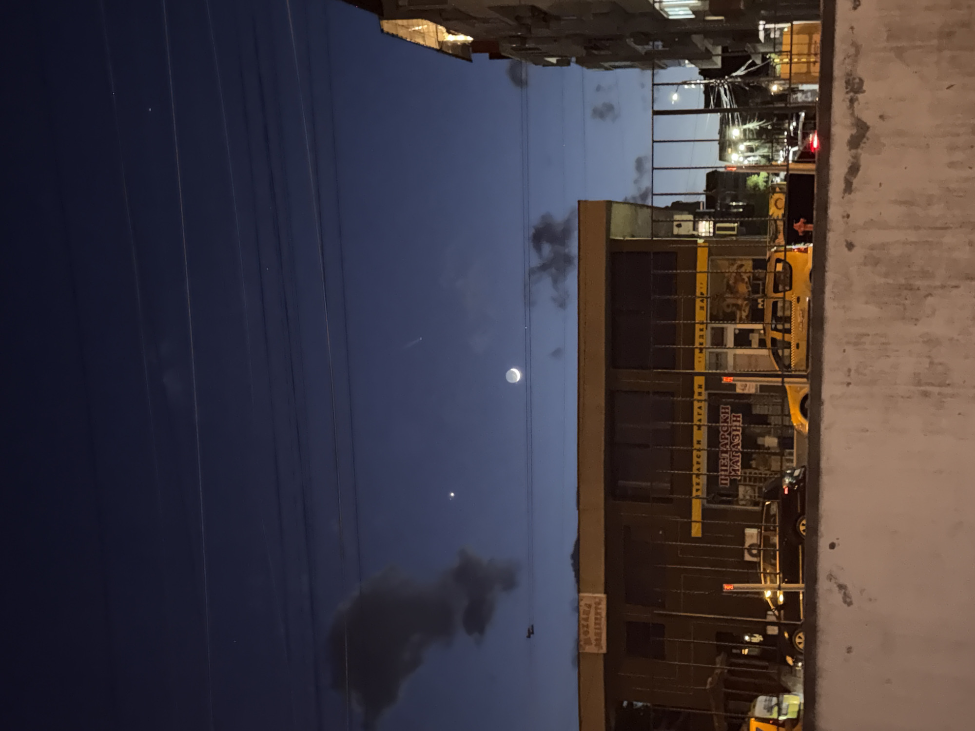 View of the night sky with a partial moon and a few stars visible above a building with lit windows. Power lines are strung across the sky. The building is a "Bulgarian Shop." The image appears to be taken from a higher location, possibly a window or balcony, looking out onto the street. The right side of the photo shows a blurred wall or structure. The location is in Burgas, Bulgaria. There are cars parked on the street.