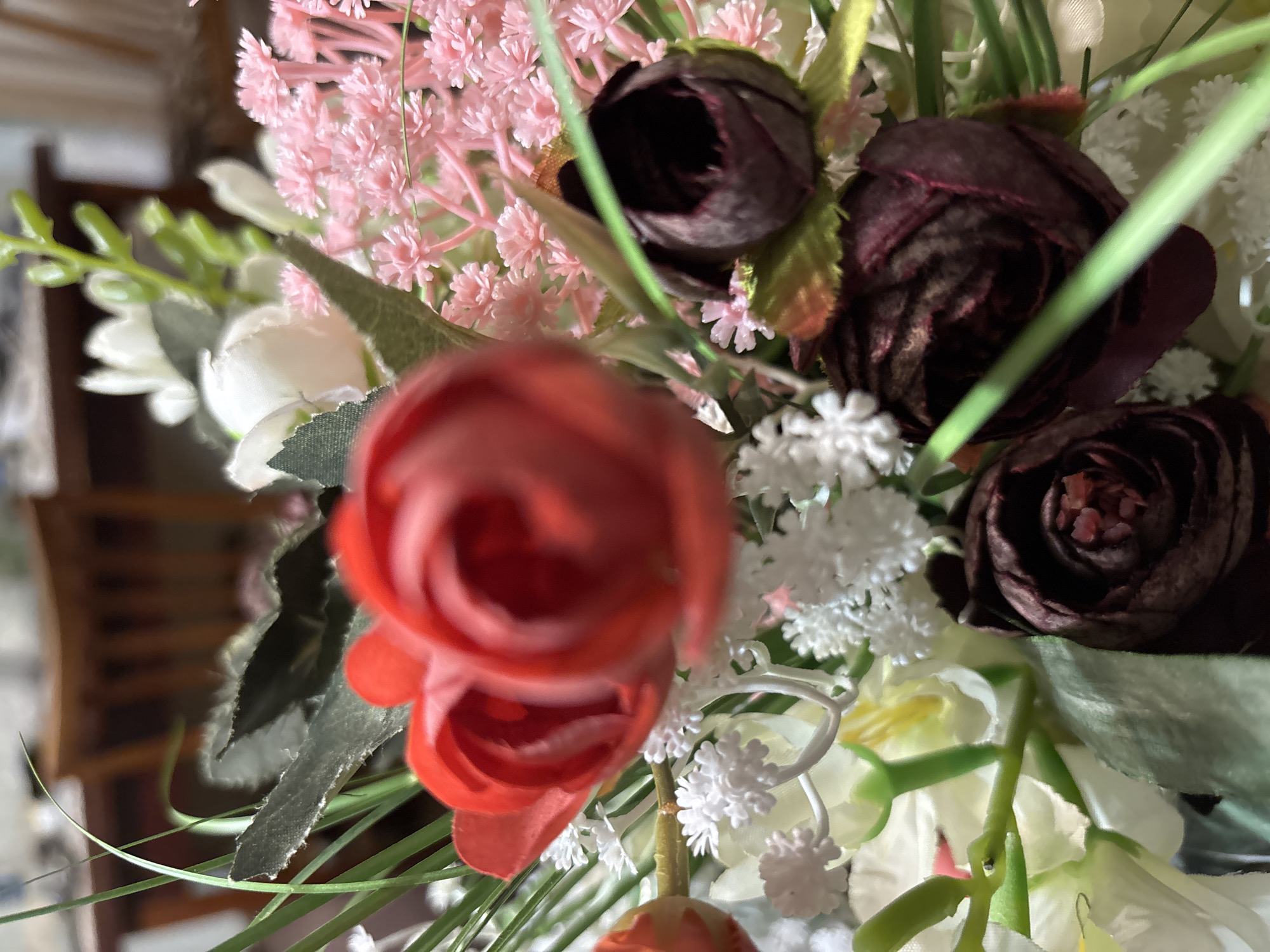The image presents a close-up, high-angle shot of an artificial floral arrangement, filling the majority of the frame. The primary subjects are various types and colors of faux flowers and greenery.
In the immediate foreground, a vibrant red rose or similar flower is significantly out of focus, appearing as a blurred swirl of red petals and providing a sense of depth. Further into the arrangement, several dark burgundy roses are visible, some partially open and others tightly budded, exhibiting slightly more detail and texture in their petals.
Scattered throughout the arrangement are clusters of smaller, lighter-colored filler flowers. To the left and in the background, numerous delicate pink flowers with a feathery or clustered appearance add softness. Various white filler flowers, some with star-like or snowflake-like shapes, are interspersed, particularly on the right side and towards the bottom. Hints of larger white or cream-colored flowers with yellow centers can also be seen at the bottom right.
Lush green leaves and stems of varying shades and textures are woven throughout the bouquet, providing a natural contrast to the blossoms. Some leaves appear slightly crinkled, reinforcing the artificial nature of the display.
The background is heavily blurred, suggesting a shallow depth of field. In some areas, particularly behind the red foreground flower, indistinct shapes and colors imply an indoor setting, possibly showing parts of wooden furniture like a chair or table. No specific activity is depicted, as the image is a still life. There is no visible text present in the image. The overall impression is one of decorative beauty within an indoor environment.