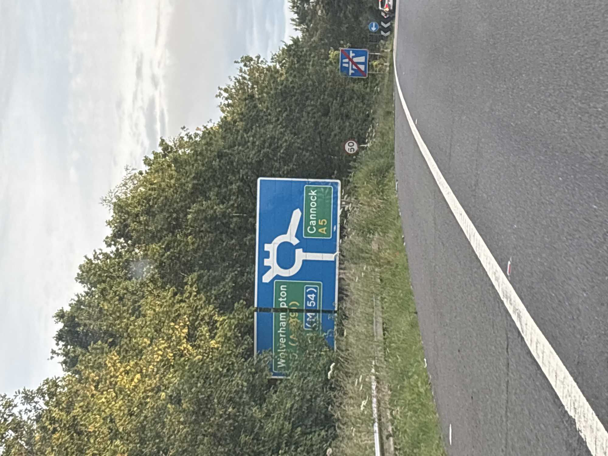 A road sign indicates directions to Wolverhampton and Cannock. The sign, which is blue and white, shows a diagram of a roundabout with arrows pointing to the destinations. "Wolverhampton" and "A5" are visible on the left side of the sign, along with "(M 54)" in a green box. On the right side, "Cannock" and "A5" are displayed. The sign is positioned next to a road in a rural or semi-rural setting, with trees and vegetation on the left and a clear sky above. A circular speed limit sign with "50" is visible below the main road sign. Further down the road, another sign indicating the end of a dual carriageway is present. A single white line marks the edge of the road. The scene is captured from the perspective of someone on the road, suggesting a driving context. The location is South Staffordshire, United Kingdom.