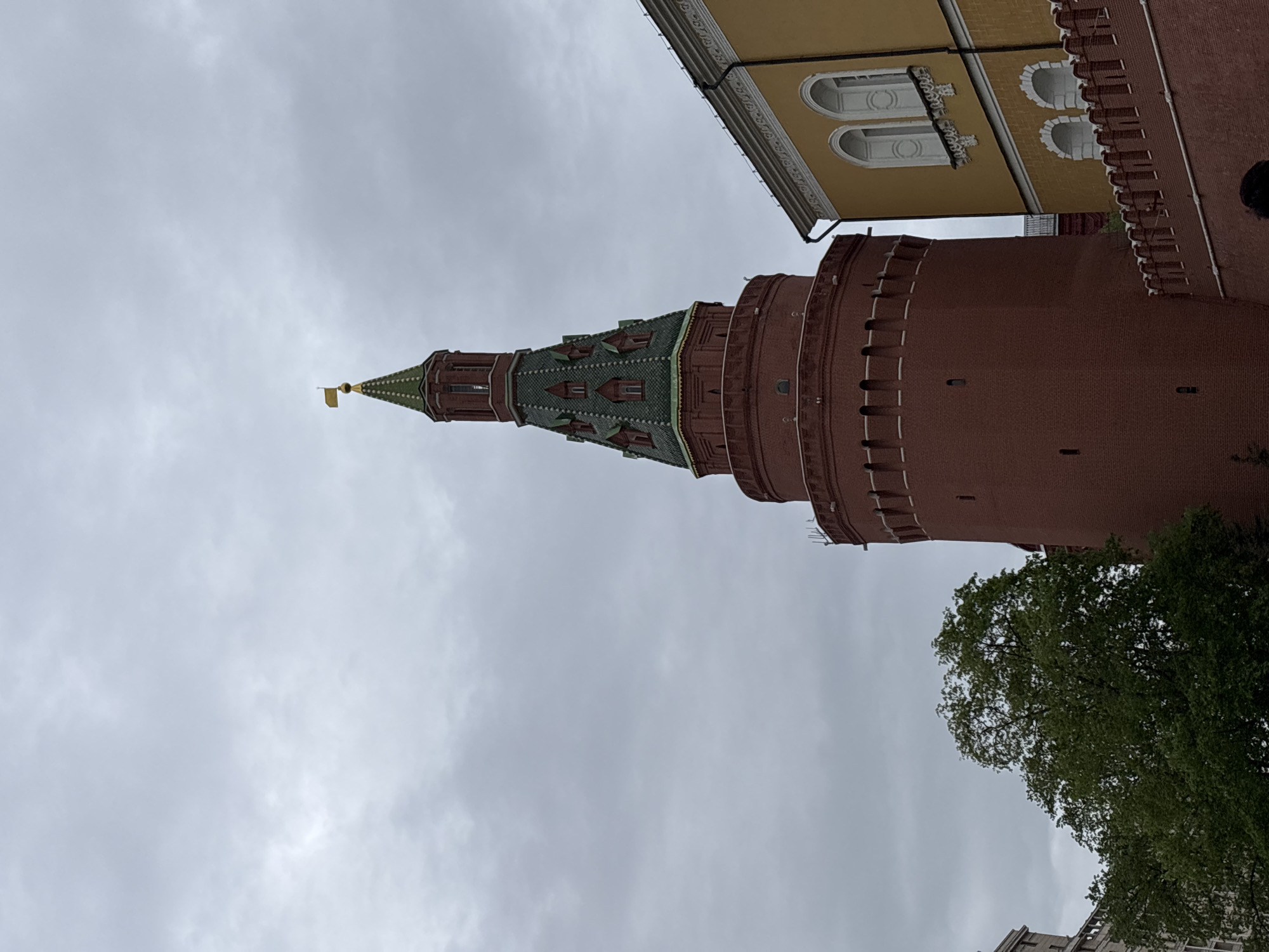 The image features a view of the Kremlin in Moscow, Russia. A red brick tower with a green-roofed spire topped with a golden flag stands prominently against a cloudy sky. A yellow building with arched windows is visible to the side of the tower. A tree with green leaves is situated near the base of the tower.
