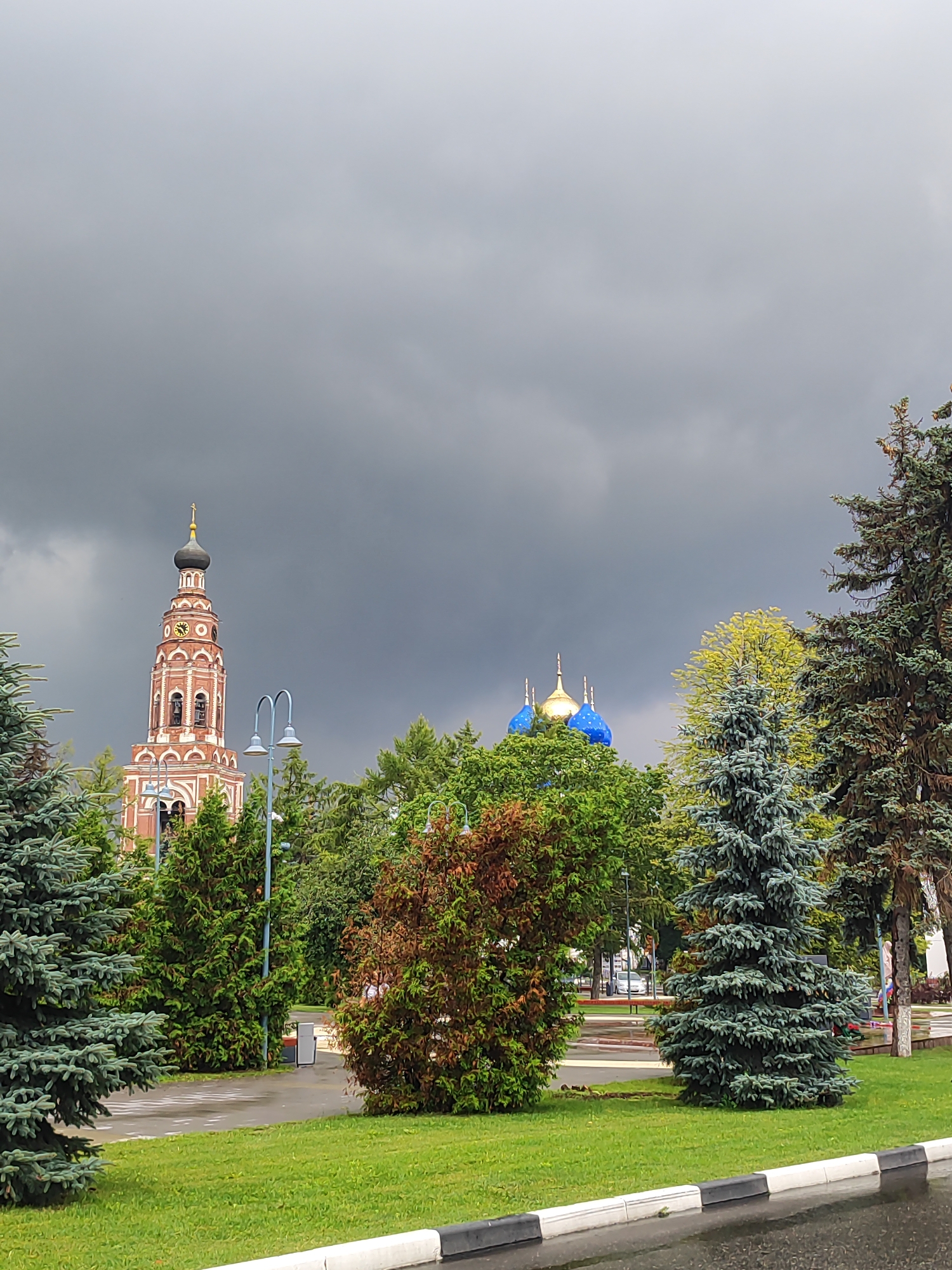 The image shows a view of the Ascension Cathedral in Bronnitsy, Russia. The cathedral's bell tower, with its red brick and white detailing, is visible on the left. To the right of the bell tower, three blue domes with golden crosses are visible, belonging to another part of the cathedral complex. The sky is overcast with dark clouds. In the foreground, there is a lush green lawn and several trees, including a large blue spruce tree on the right. A wet asphalt road with a white and black curb is in the bottom right corner, suggesting recent rain. The overall scene is an exterior shot of a religious landmark within a park-like setting.