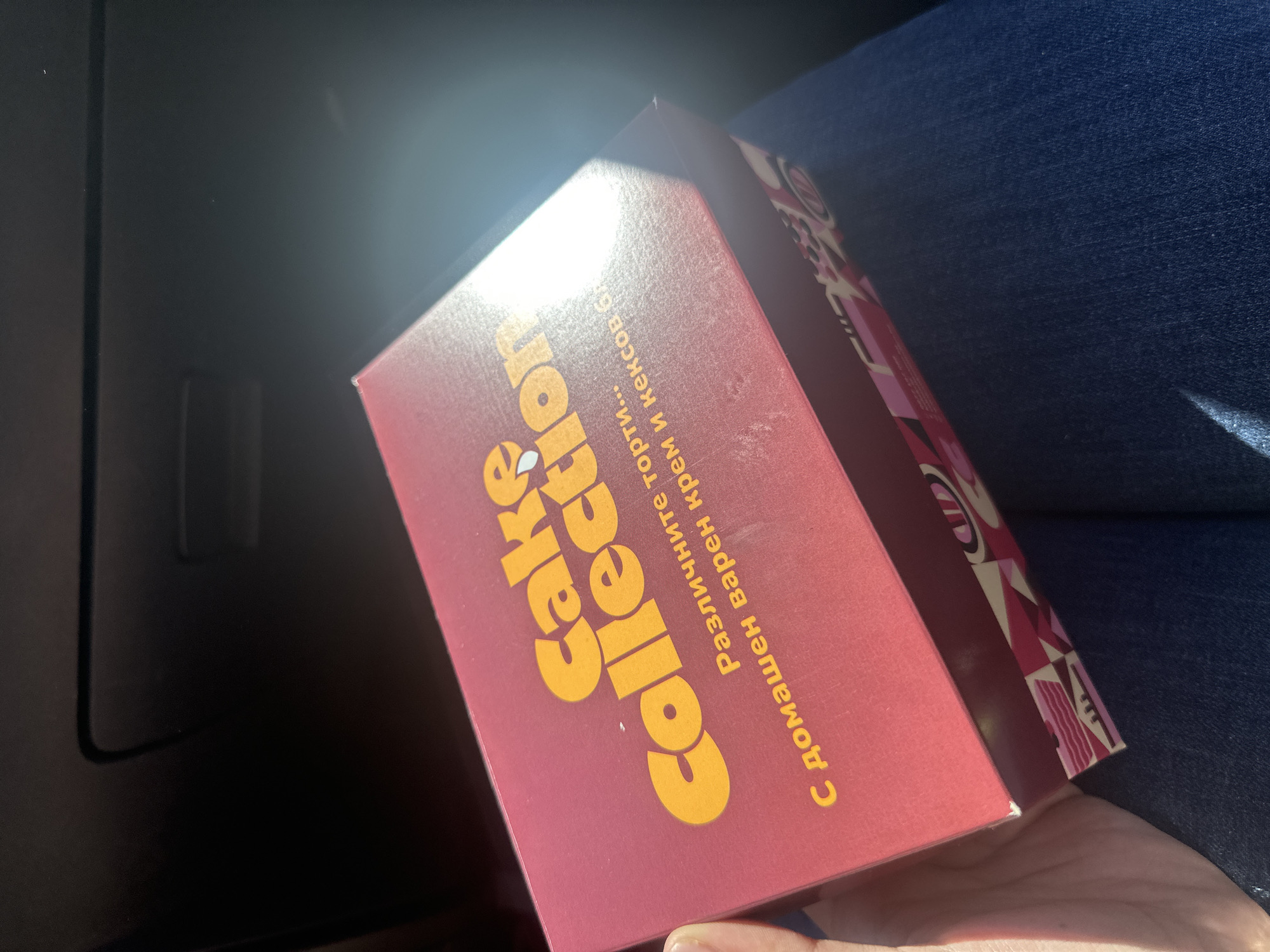 A maroon-colored box with "Cake Collection" written in yellow is being held in someone's hand. The text on the box is in Bulgarian and includes "с домашен варен крем и кексов". The box is partially in sunlight and there is a laptop and jeans in the background. The location is unknown, and therefore, impossible to state if it is in Plovdiv, Bulgaria.