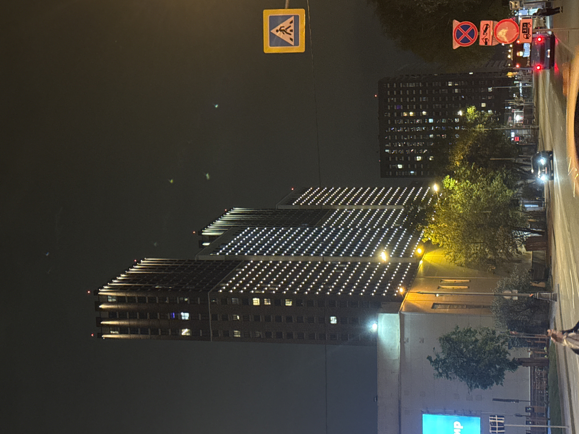 Night scene in Moscow, Russia, featuring high-rise buildings.
Two large buildings are visible, both illuminated with rows of white lights across their facade. A street runs along the right side of the image. Streetlights illuminate trees and the road, and there are visible road signs. A pedestrian crossing sign is visible on the left side of the street. Other road signs are seen in the right side of the image. The sky is dark.