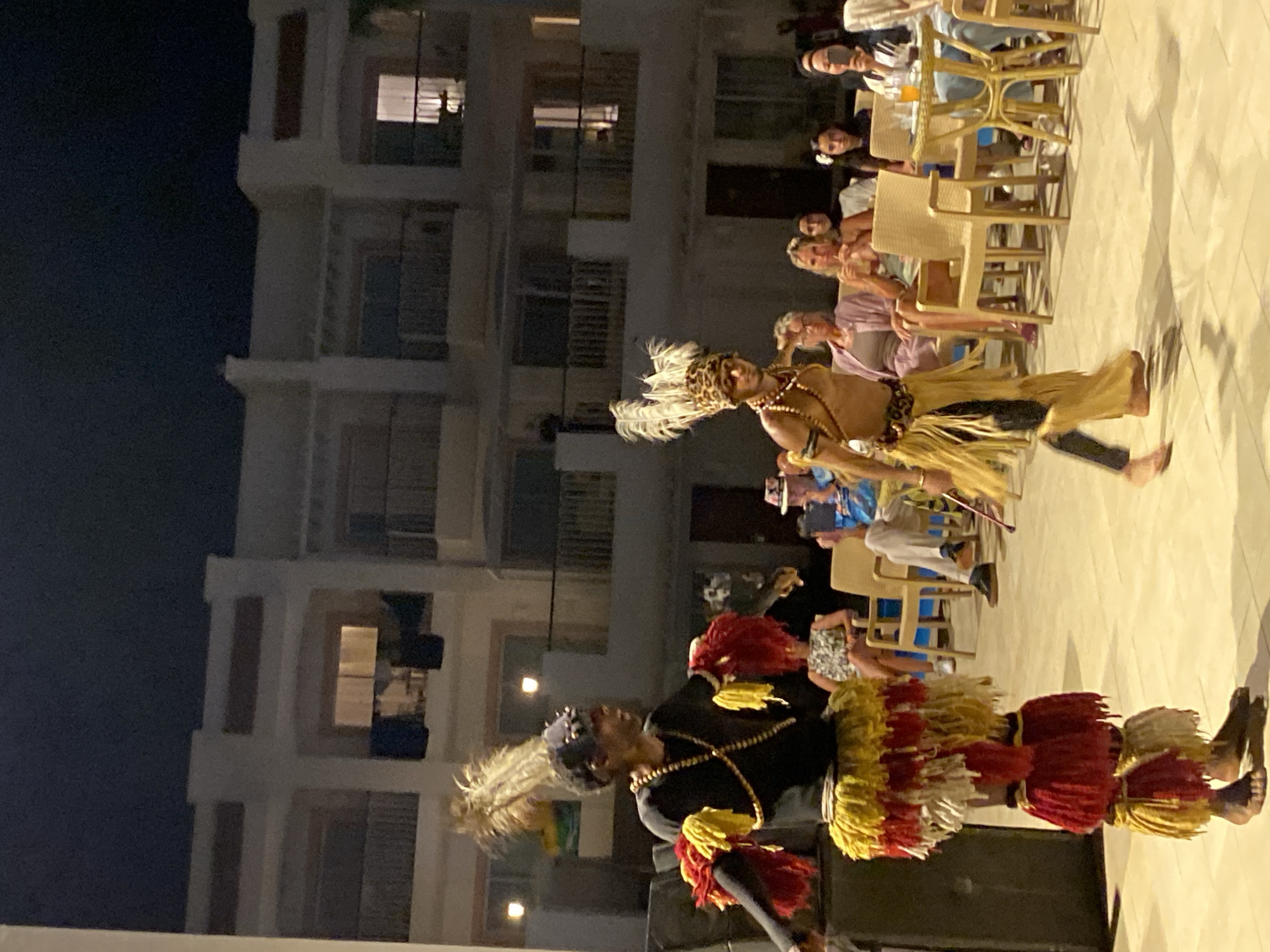 Two male performers in traditional African attire are dancing outdoors at night in Sharm El Sheikh, Egypt. The performer on the left wears a black long-sleeved shirt, a beaded necklace, a headdress with feathers, and a skirt made of layered red, yellow, and white tassels. He also has red and yellow fringe on his arm coverings. The performer on the right is shirtless and wears a headdress with feathers and what appears to be a leopard-print band, a beaded necklace, and a skirt made of yellow fringe. They are on a tiled outdoor surface in front of a multi-story building with balconies and lit windows. Several people are seated at tables in the background, observing the performance. The overall setting suggests a resort or hotel complex.