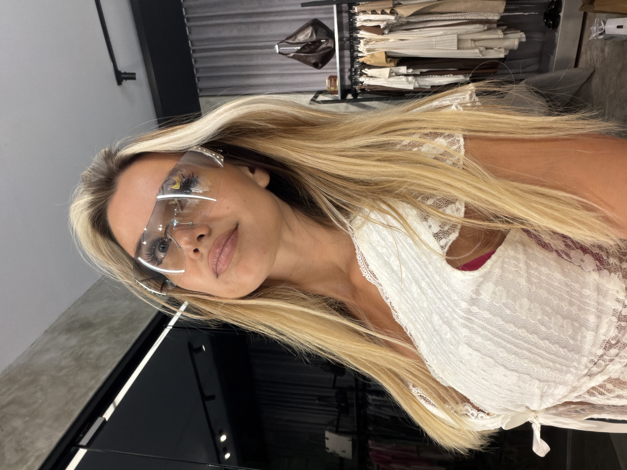 A blonde woman wearing a white lace dress and large futuristic sunglasses poses for a mirror selfie in what appears to be a boutique clothing store. The store is visible in the background, with racks of clothing and a dark leather handbag on a shelf. The woman's makeup is visible, with bold eyelashes and lip gloss. The scene suggests a personal moment of self-expression and style experimentation within a retail environment.