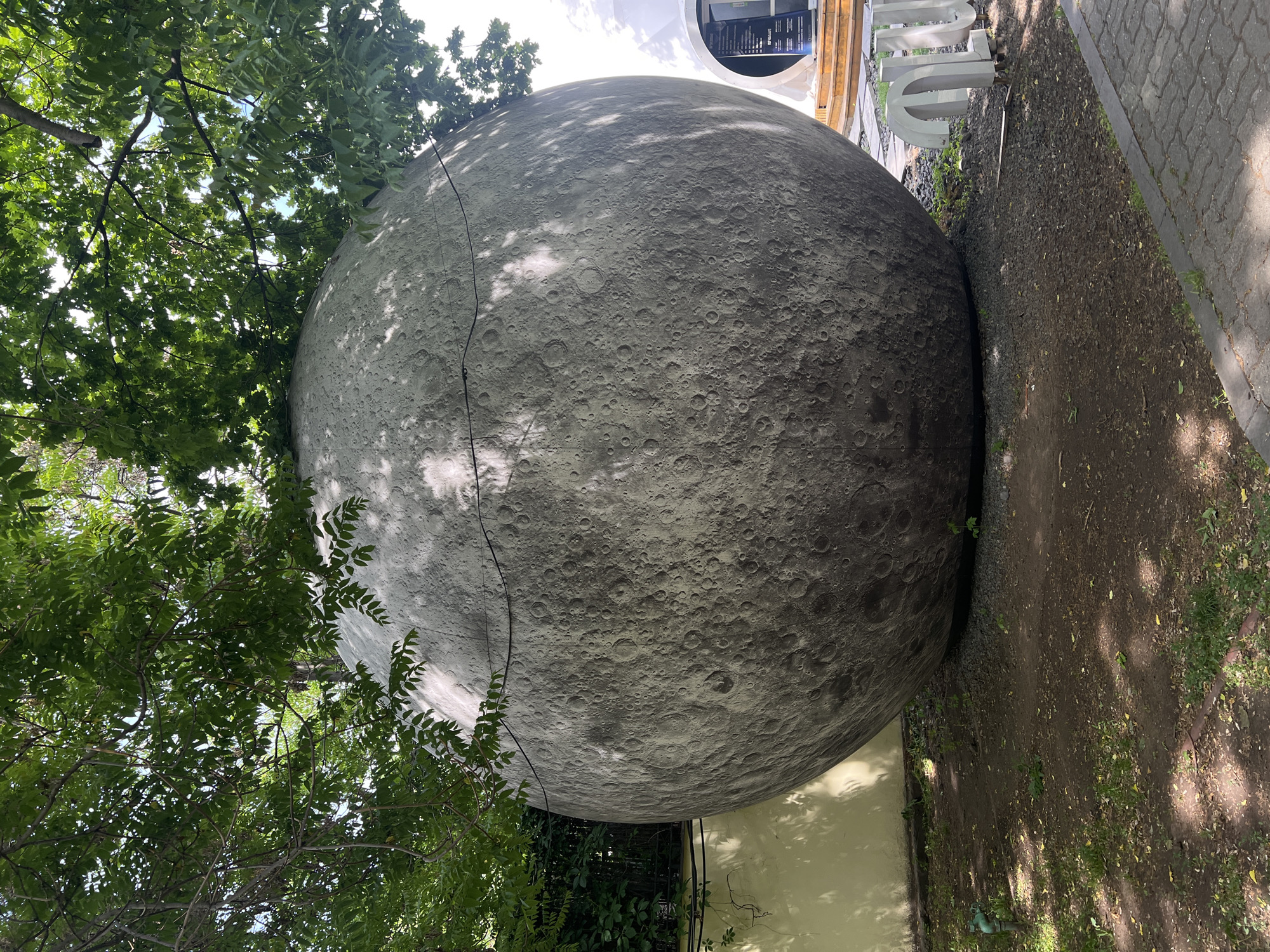The image depicts a large-scale model of the moon, placed outdoors adjacent to trees and a building. The moon model has a textured surface representing lunar craters and is bisected by a crack running across its surface. To the left of the moon model are dense green trees with visible branches and leaves. To the right is a pathway covered with brown soil, bordered by paving stones. Behind the moon, parts of a building are visible, including white architectural elements, possibly letters. The photograph is taken at a slight upward angle, and the location is given as Unknown, Unknown.