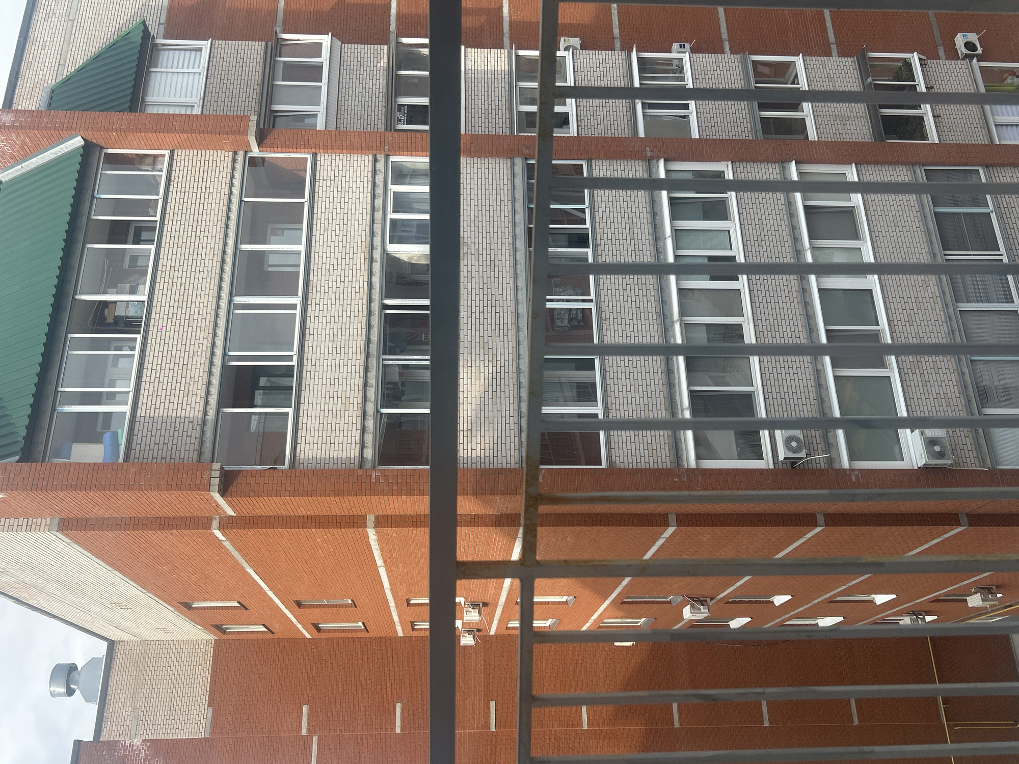 The image shows a brick building viewed through a metal grate. The building has a combination of red and light-colored brick, and rows of windows. Some windows are open. Some windows have air conditioning units installed below them. The top left of the building has a green awning. The image appears to be taken from an elevated position looking down at the building. Based on the reference images, this is located in Chaikovsky, Russia.