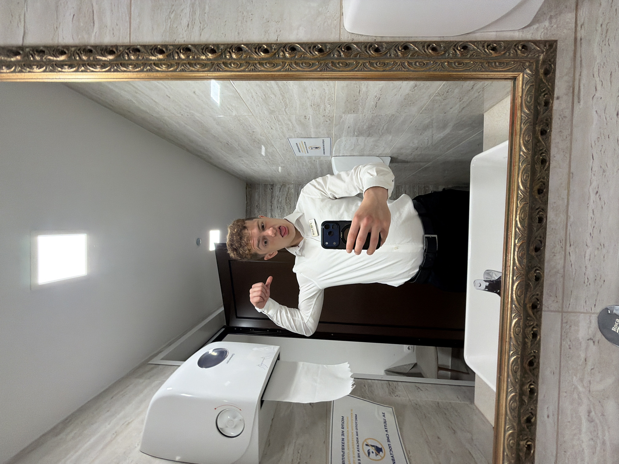 A young man is taking a selfie in a bathroom mirror in Varna, Bulgaria. He is wearing a white collared shirt, black pants, and a name tag. He has curly blonde hair and is sticking his tongue out. He is holding a black smartphone in his left hand and giving a thumbs up with his right hand.
The bathroom has light-colored marble walls and tile floors. There is a toilet, a sink, a paper towel dispenser, and a sign on the wall. The mirror has a gold ornate frame. A bright square light fixture is visible on the wall.