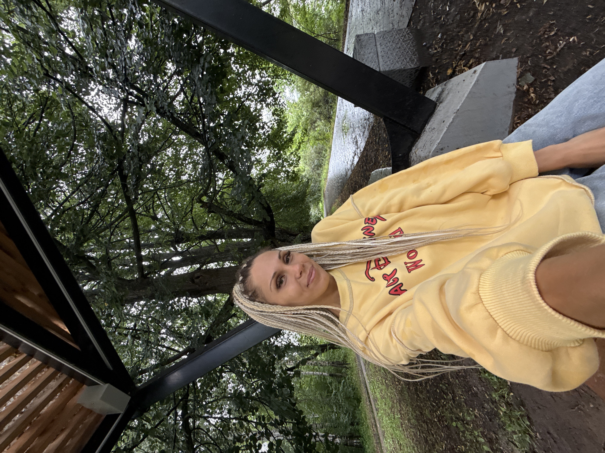 In Rusanovo, Russia, a young woman with blonde braided hair is taking a selfie in a park. She is wearing a yellow sweatshirt with red writing. The woman is sitting on a concrete bench under a wooden and metal canopy. Trees and foliage surround the area, and a wet paved path is visible in the background. The lighting suggests an overcast day.