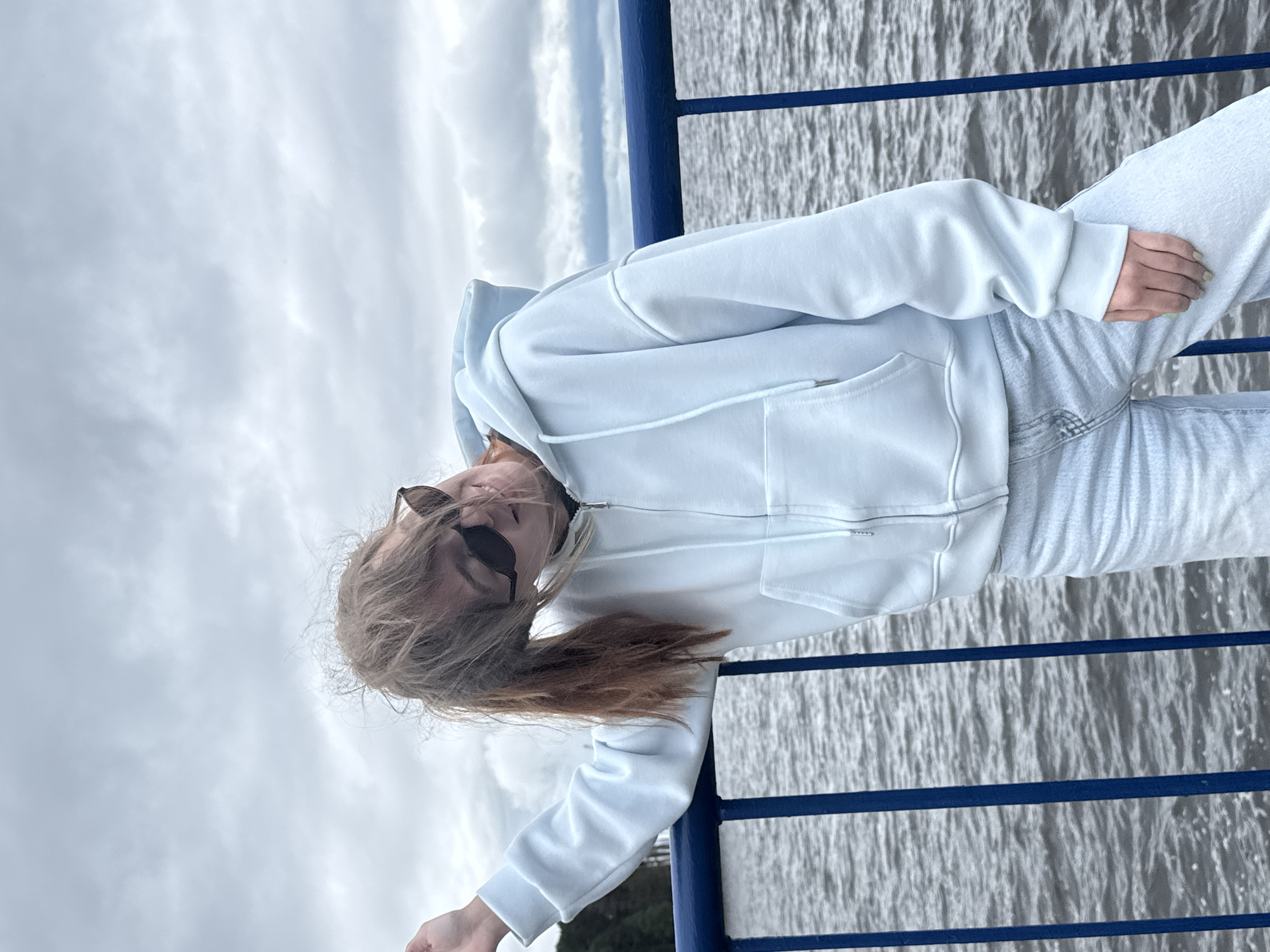 An outdoor, eye-level full-shot depicts a young individual leaning on a blue metal railing beside a body of water in Chaikovsky, Russia, under an overcast sky. The subject, a female with light brown, wind-blown hair pulled into a low ponytail, is wearing black sunglasses, a light blue full-zip hoodie, and light blue denim jeans. Her head is sharply tilted to her left (viewer's right), and she displays a slight smile. Her left forearm rests on the top bar of the blue railing, which features multiple vertical bars. The background consists of dark, rippling water and an expanse of grey, cloudy sky. A faint, distant treeline is visible on the horizon to the left.