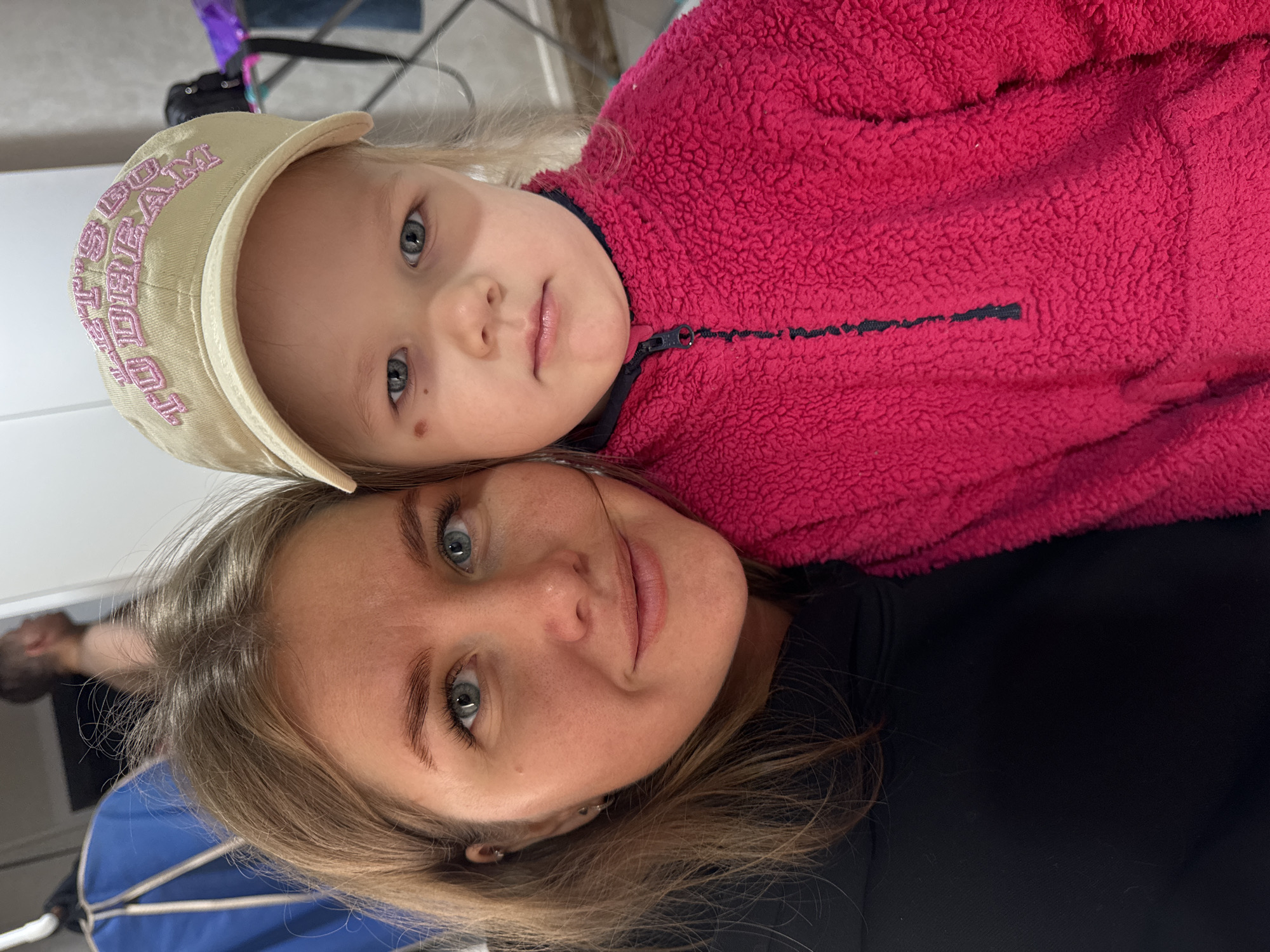 The image shows a woman and a young girl, seemingly in a close, possibly familial, relationship. The woman is wearing a black top and has blonde hair. The young girl is wearing a pink fleece jacket and a light-colored baseball cap with pink lettering. They are posed closely together, appearing to be taking a selfie or having someone else take their picture. The background is out of focus and indistinct, but it appears to be indoors. It is unknown if the location is in the city of Ферма, Russia.