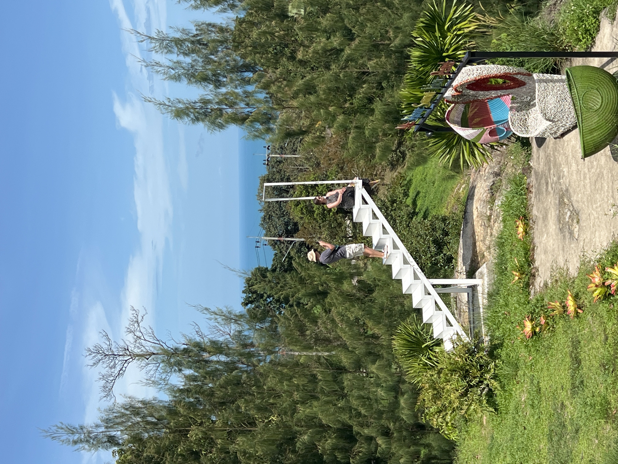 This outdoor image displays a scenic viewpoint in Baan Thurian, Thailand, under a bright blue sky with sparse white clouds. In the foreground, a concrete path is bordered by green grass and several plants with reddish-yellow leaves. To the right, a black metal stand supports an artistic arrangement of three woven elements: two multi-colored (pink, blue, green, white) abstract forms positioned above a green, bowl-shaped structure. A partially obscured sign with multi-colored lettering is visible above the stand.
Centrally located is a white, cantilevered staircase structure. A fair-skinned male, wearing a light hat, dark grey t-shirt, and light shorts, stands on a lower step, holding a dark smartphone to photograph. He is facing towards the upper section of the staircase. On the topmost step, a fair-skinned female, wearing dark sunglasses, a dark patterned top, and dark lower garments, is seated with her legs dangling, looking towards the left.
The mid-ground and background feature dense clusters of tall, slender green trees. Beyond these trees, a clear blue sea extends to the horizon, visible between the foliage. The terrain appears verdant and undulating.