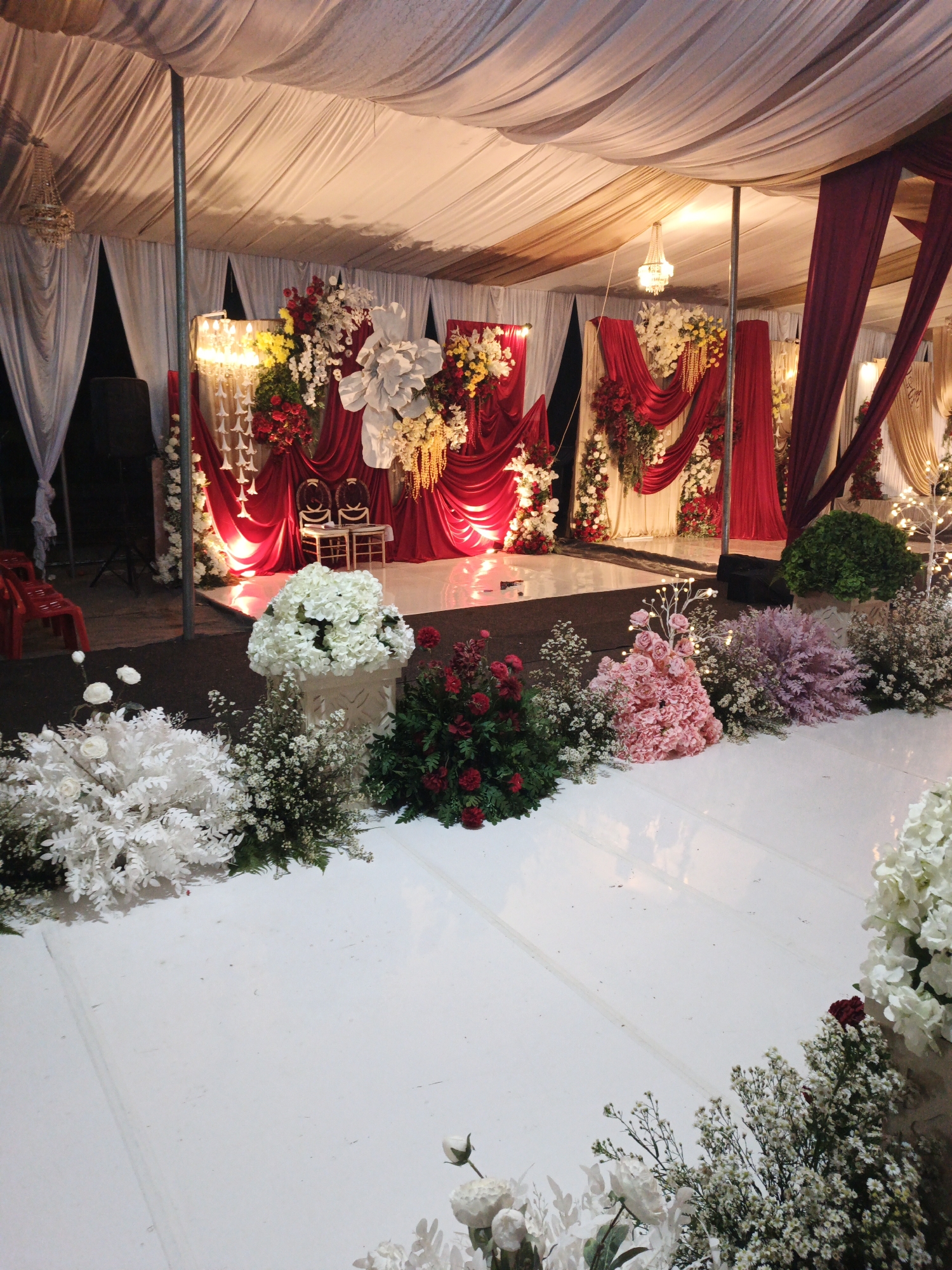 The image depicts a decorated outdoor event space, likely for a wedding or celebration, situated in Keudealue Ie Puteh, Indonesia. A white tent with draped fabric provides overhead coverage. The scene features a raised white platform, possibly a stage, adorned with elaborate floral arrangements in shades of red, white, yellow, and pink. Red and white curtains are used as a backdrop, with two chairs placed centrally on the platform. Various types of flowers, including roses and hydrangeas, are arranged in bouquets and as accents along the edges of the platform and surrounding areas. Lighting fixtures, including chandeliers, are visible, contributing to the ambiance. Red plastic chairs are present in the background, along with audio equipment. The ground surface appears to be a dark, possibly asphalt or dirt, material contrasting with the white platform. The overall impression is one of a formally set up event venue.