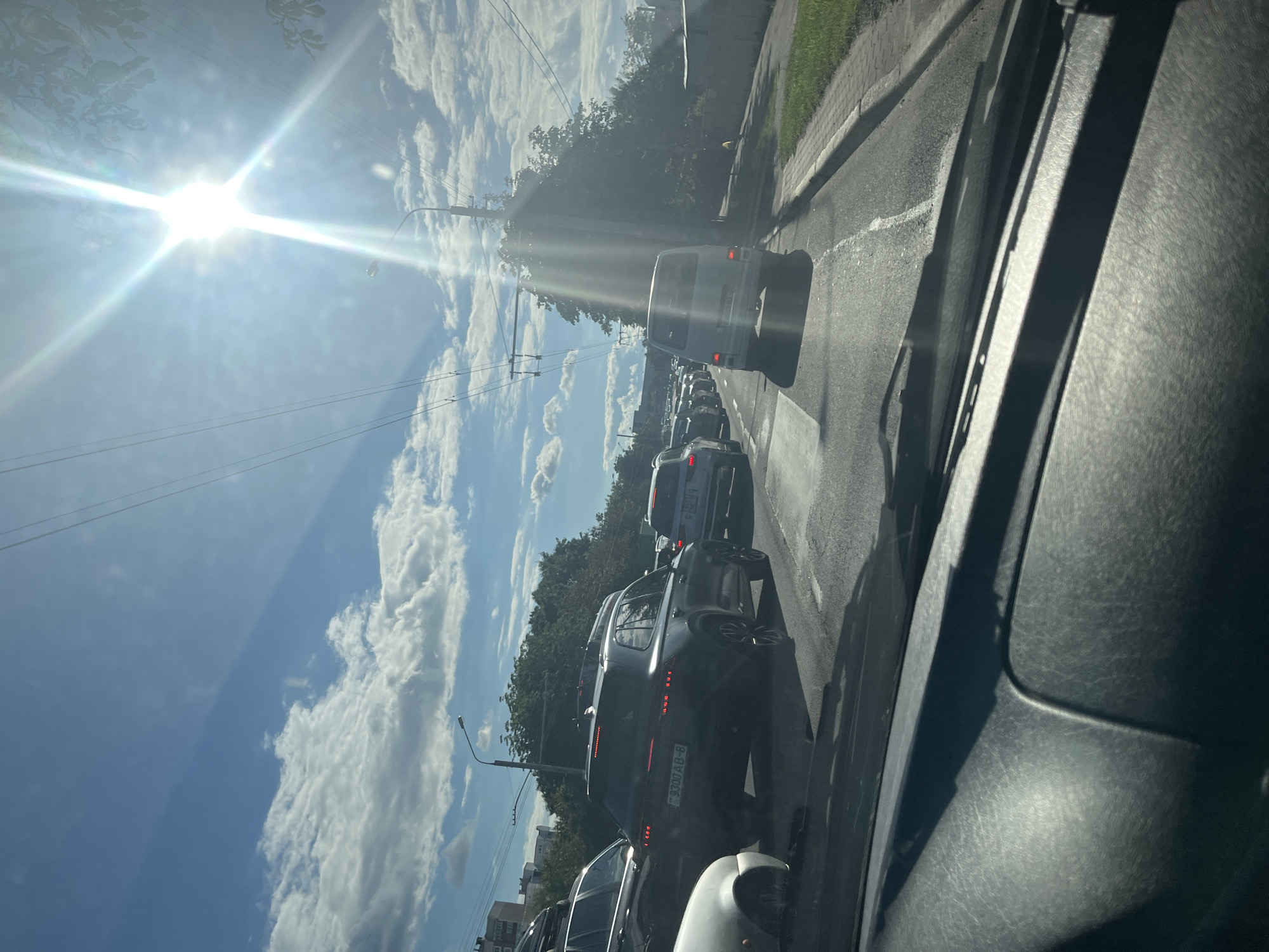 The image shows a traffic jam on a sunny day in Minsk, Belarus, viewed from inside a vehicle. Several cars are lined up on a multi-lane road, with lush green trees lining the right side of the street. Overhead power lines suggest the presence of public transportation, likely trolleybuses. The sun is bright and high in the sky, casting lens flares and creating reflections on the windshield and road. The sky is blue with scattered white clouds. Buildings are visible in the distance on the left side of the frame, indicating an urban environment. The license plate of a dark SUV in the foreground reads "9300 AB-8", which is a Belarusian license plate format.